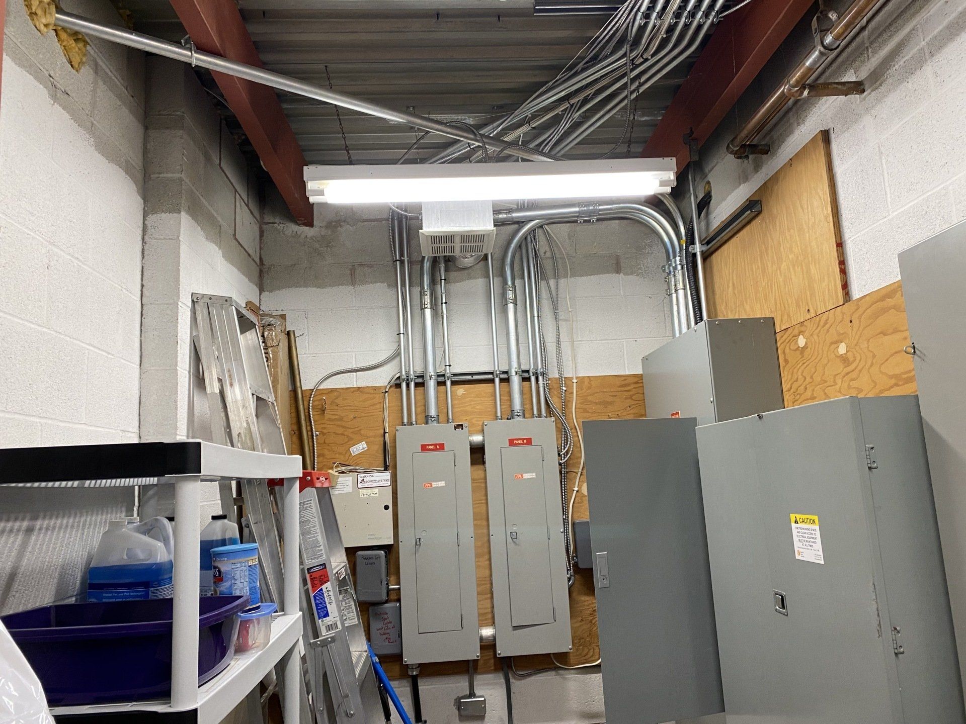 Electrical panel and wiring in a utility room. Metal conduits run to gray boxes, fluorescent light overhead.
