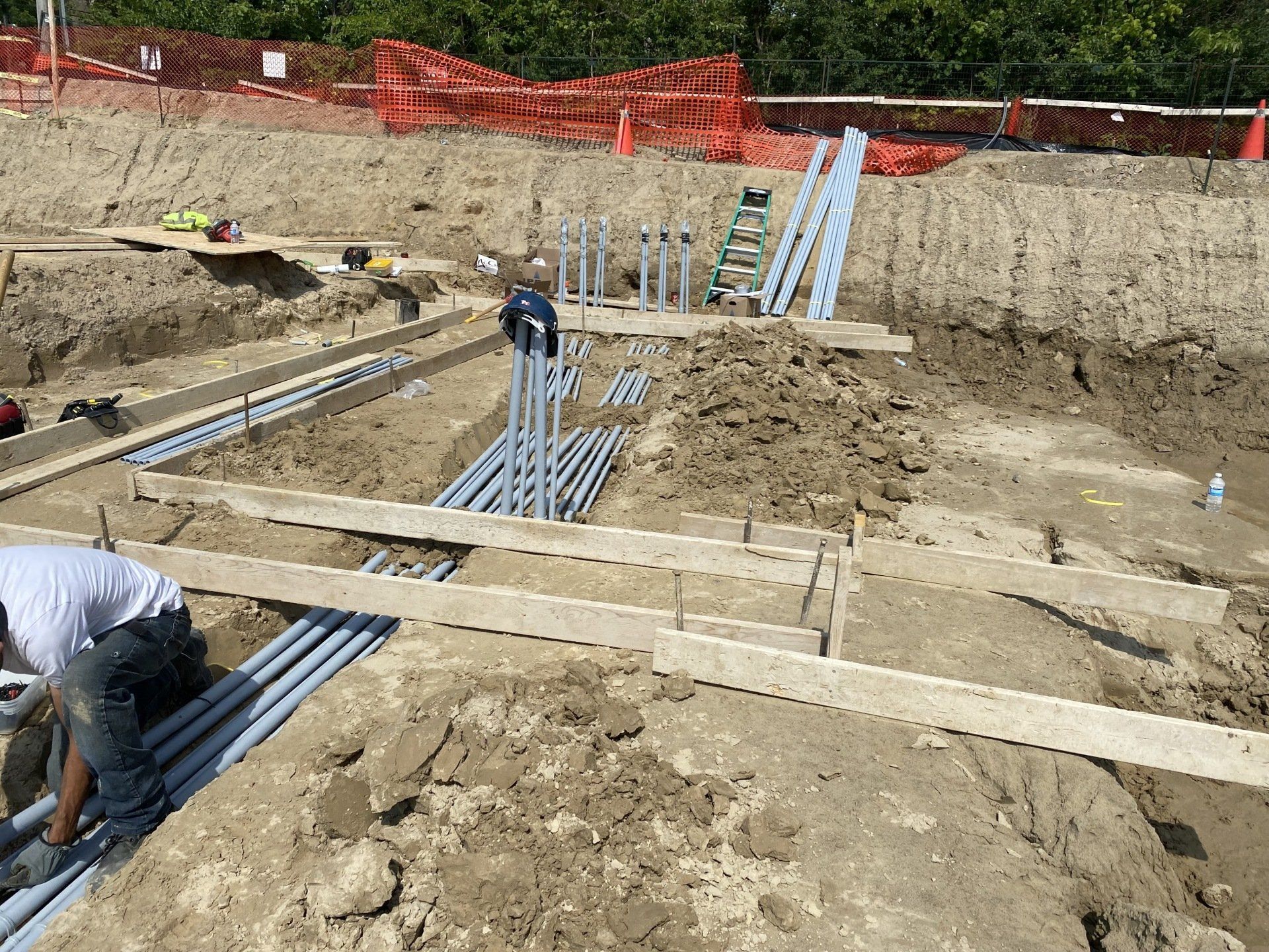 Construction workers in a dirt trench, installing conduit. Orange safety fencing and ladder present.