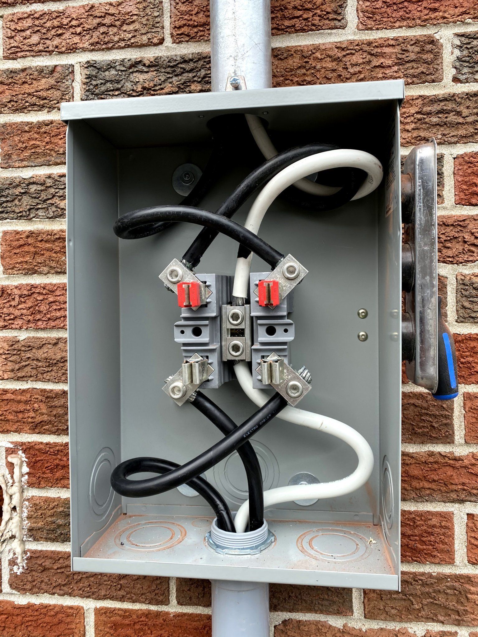 Gray electrical meter box with black and white wires against a red brick wall.