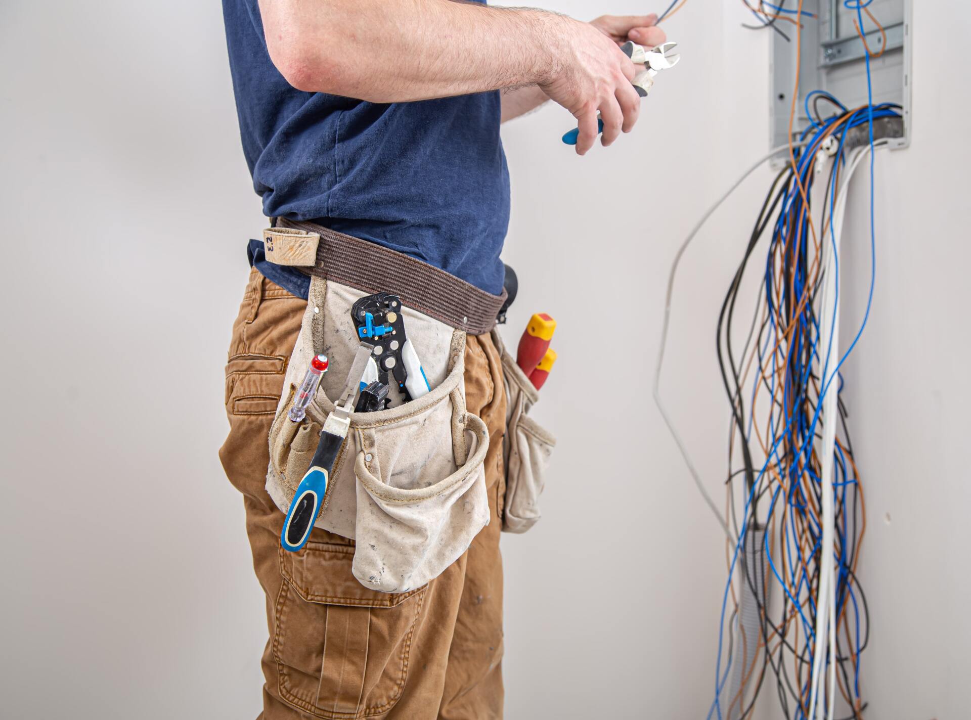 Electrician working on wiring, wearing a tool belt, near a wall with exposed wires.