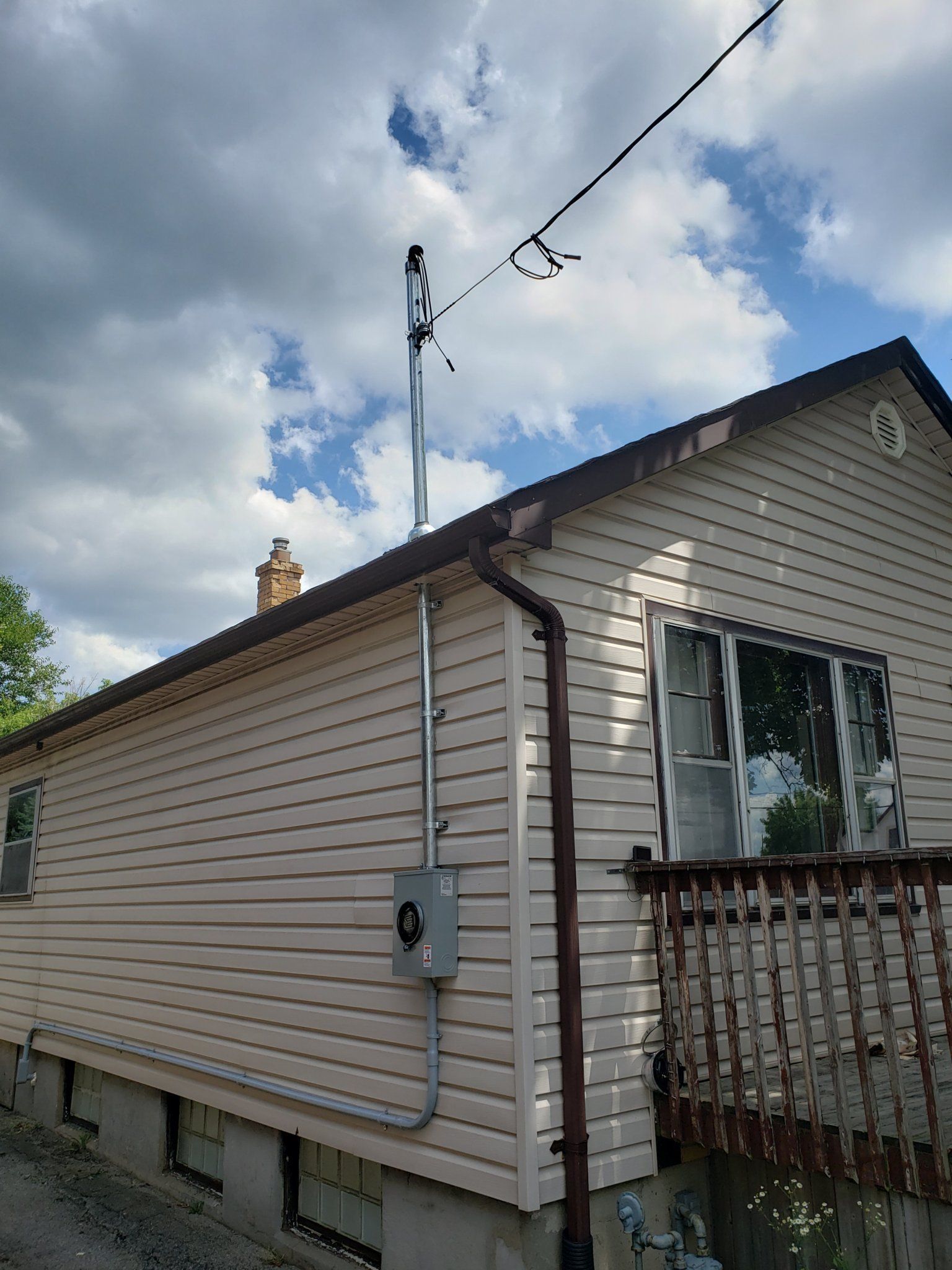 Tan house with a tall antenna and conduit on the side, a cloudy sky in the background.