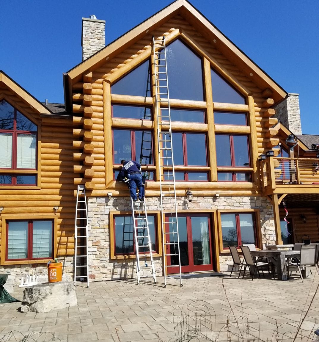 Person on a tall ladder cleaning windows of a large log cabin.