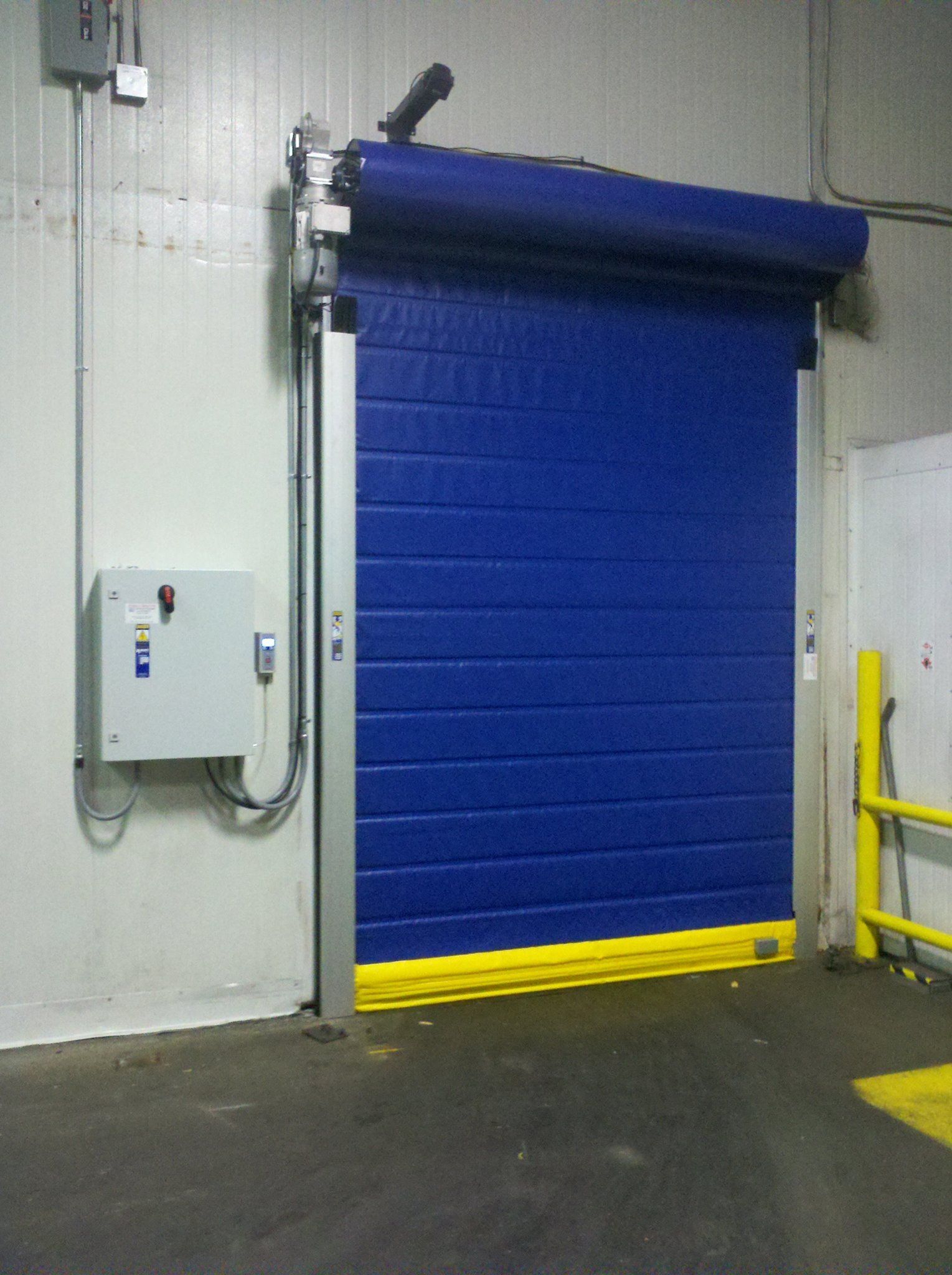 Blue industrial roll-up door, yellow base, in a concrete setting with a control box and conduit.