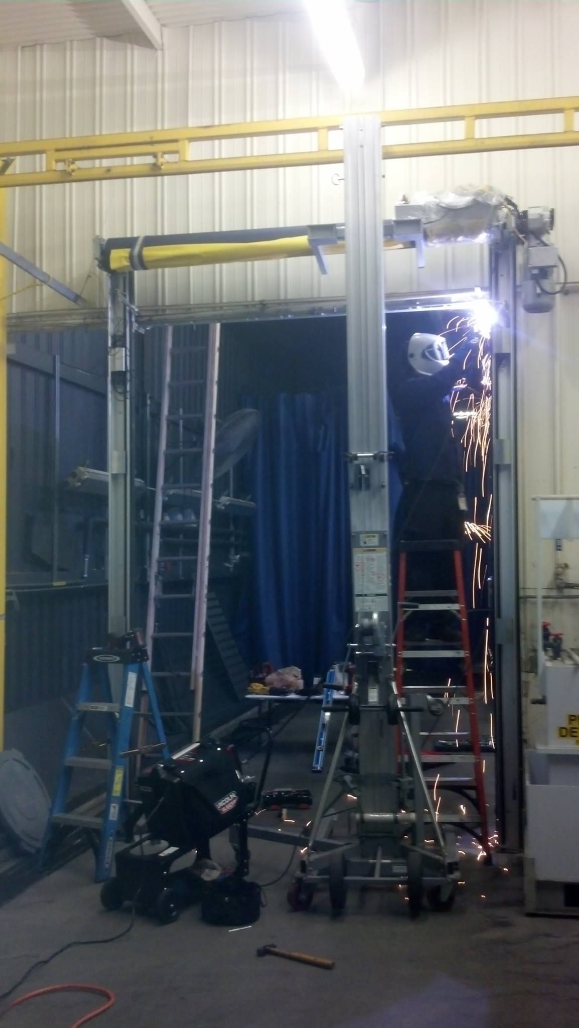 Welder working on metal frame in a factory, sparks flying. Blue curtain and ladders visible.