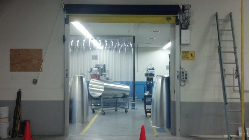 Factory interior with a large open doorway, clear plastic strips, and metal equipment on a cart.