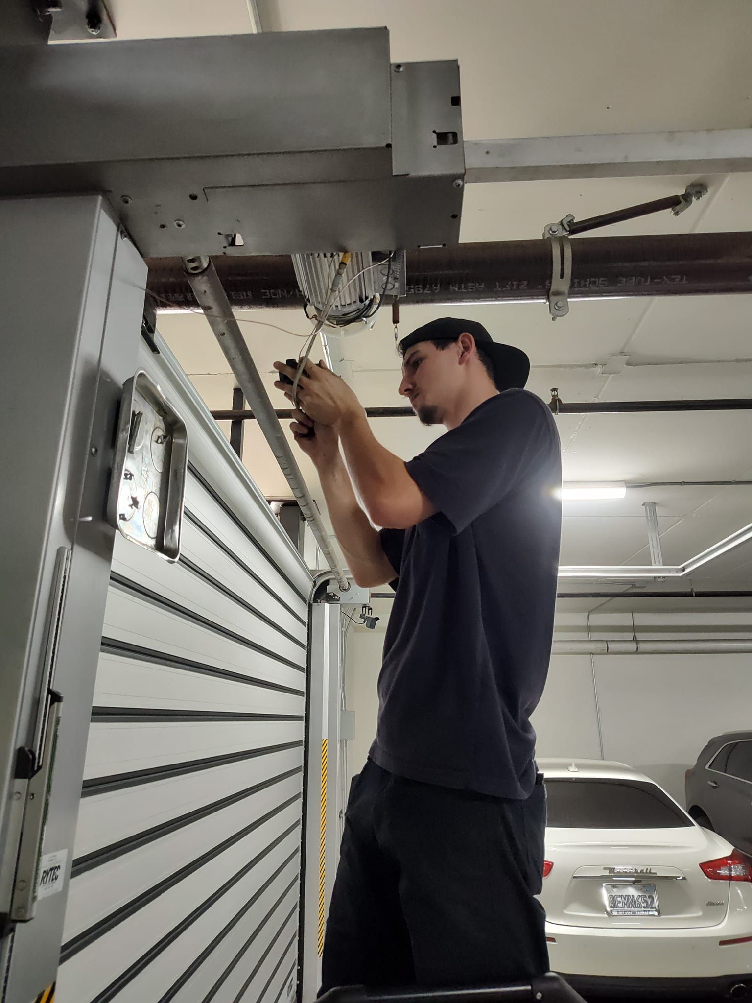 Man on a ladder repairs machinery, near a large metallic door, in a garage setting.