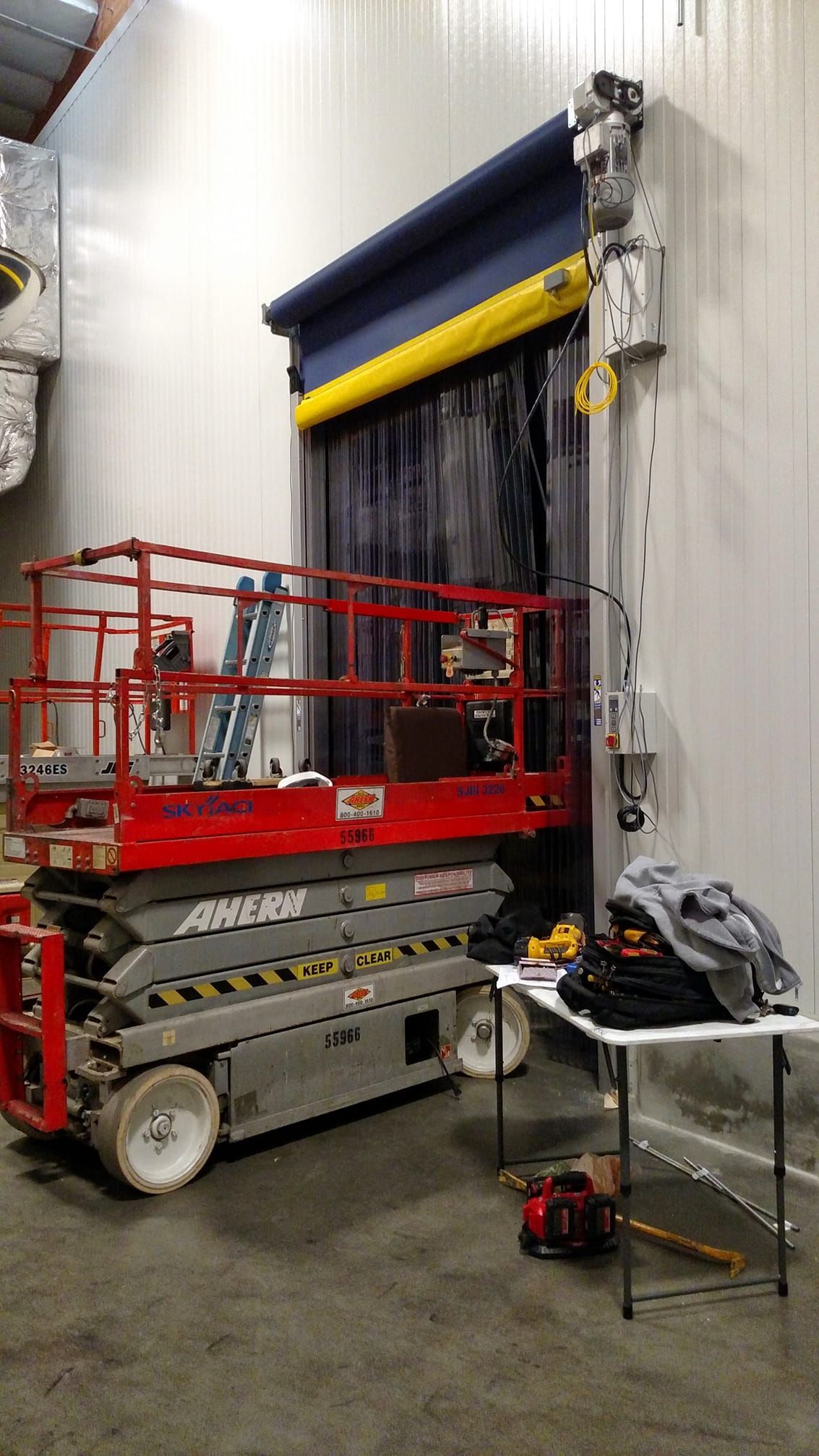 Red scissor lift near an industrial door with blue covering. A table and tools are nearby.