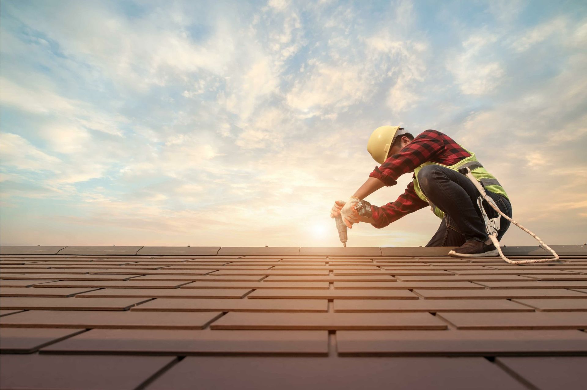 A roofer in a yellow hard hat and safety vest works on a roof with a sunset in the background