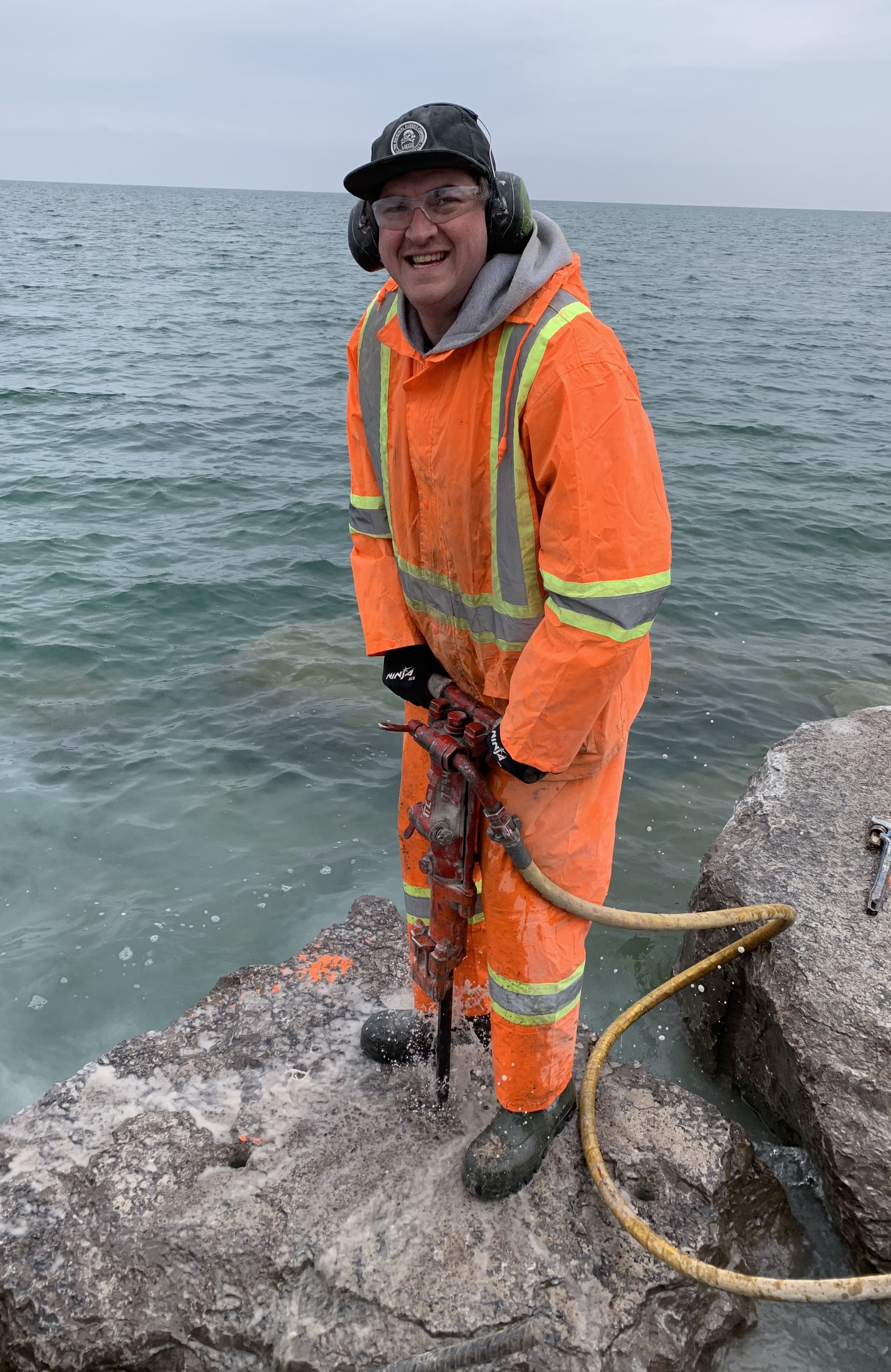 Man in orange work suit, earmuffs, drilling rock near water.