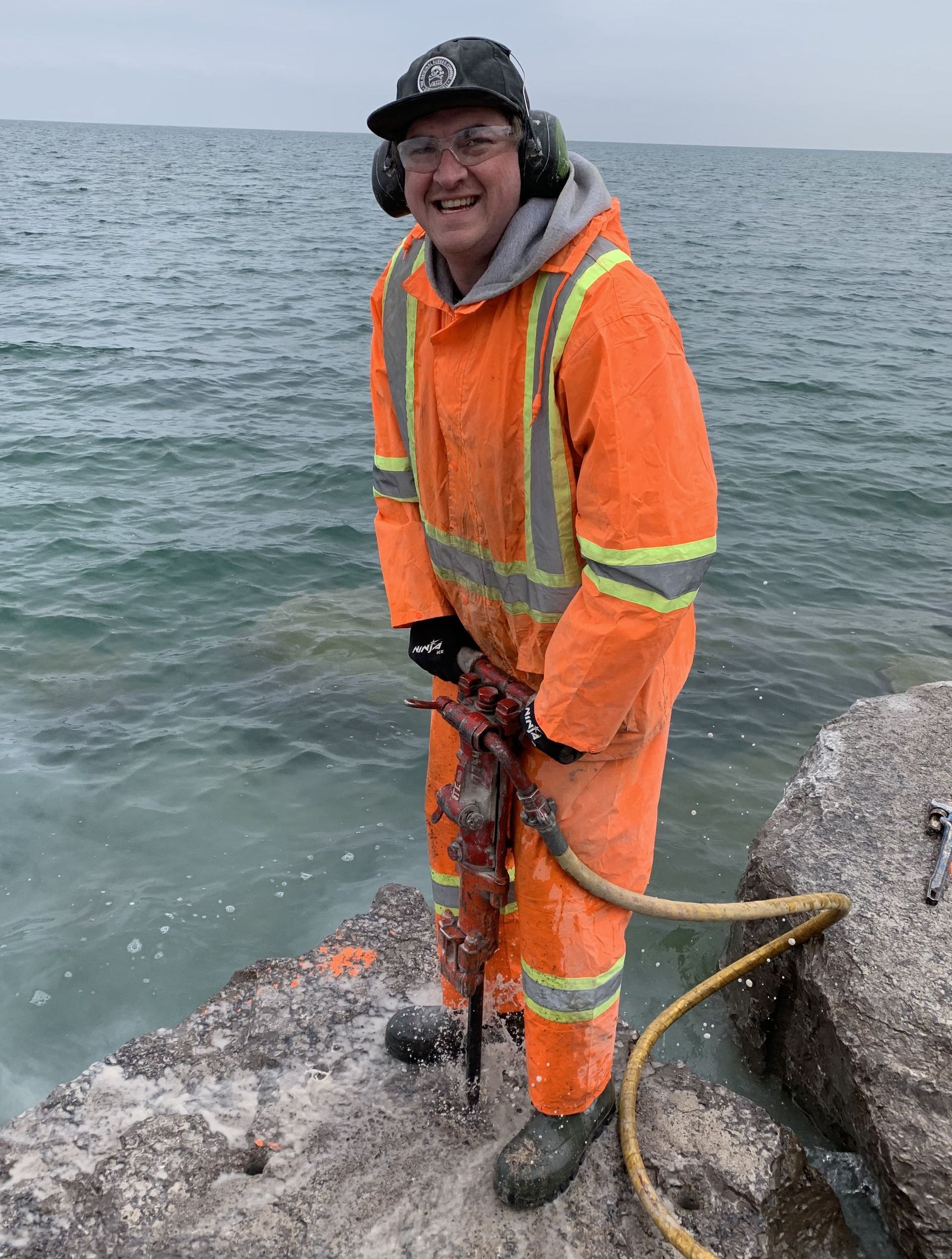 Man in orange work suit, using a jackhammer by the water.