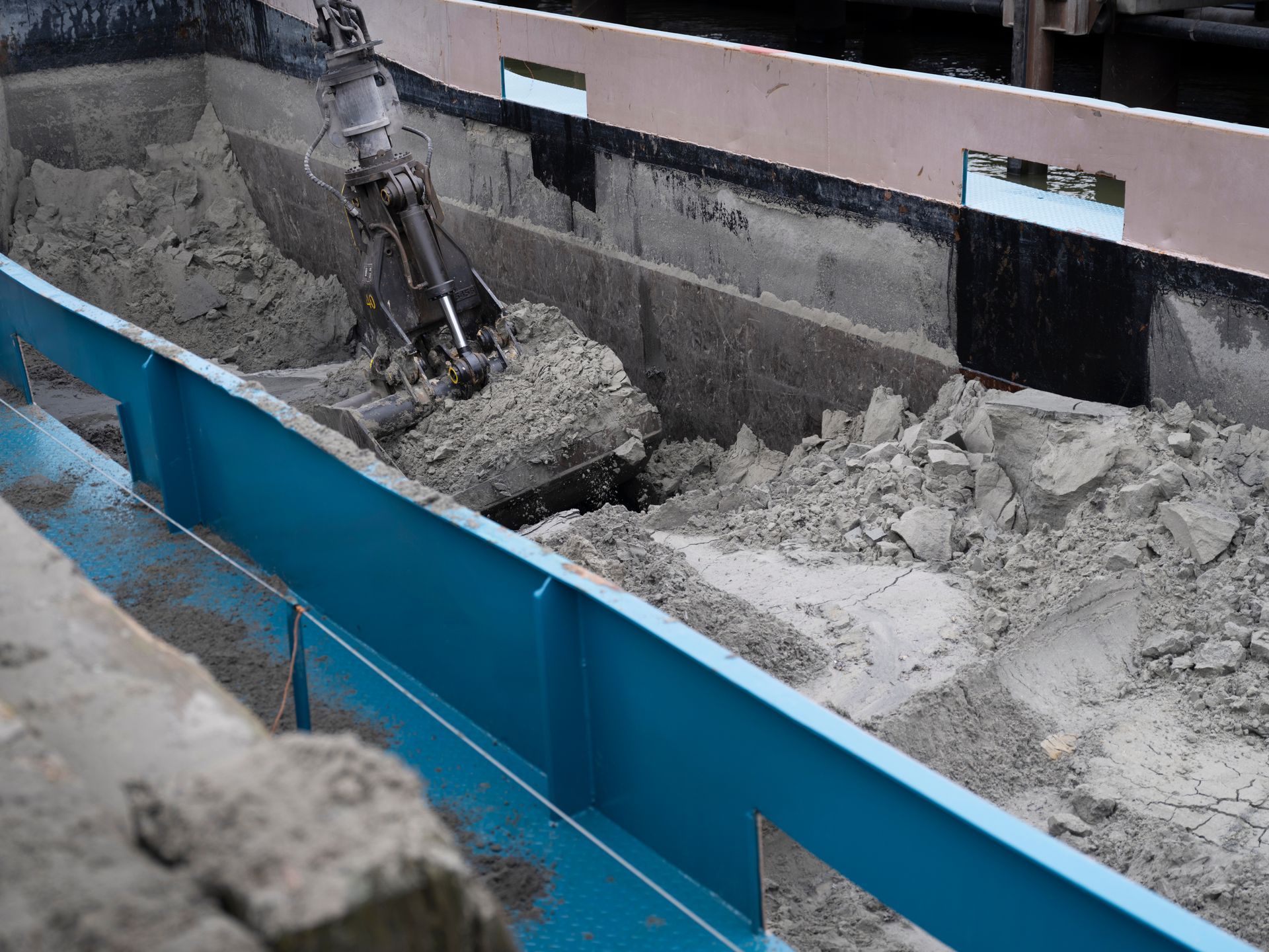 An excavator arm scoops debris from a blue-lined trough at a construction site.