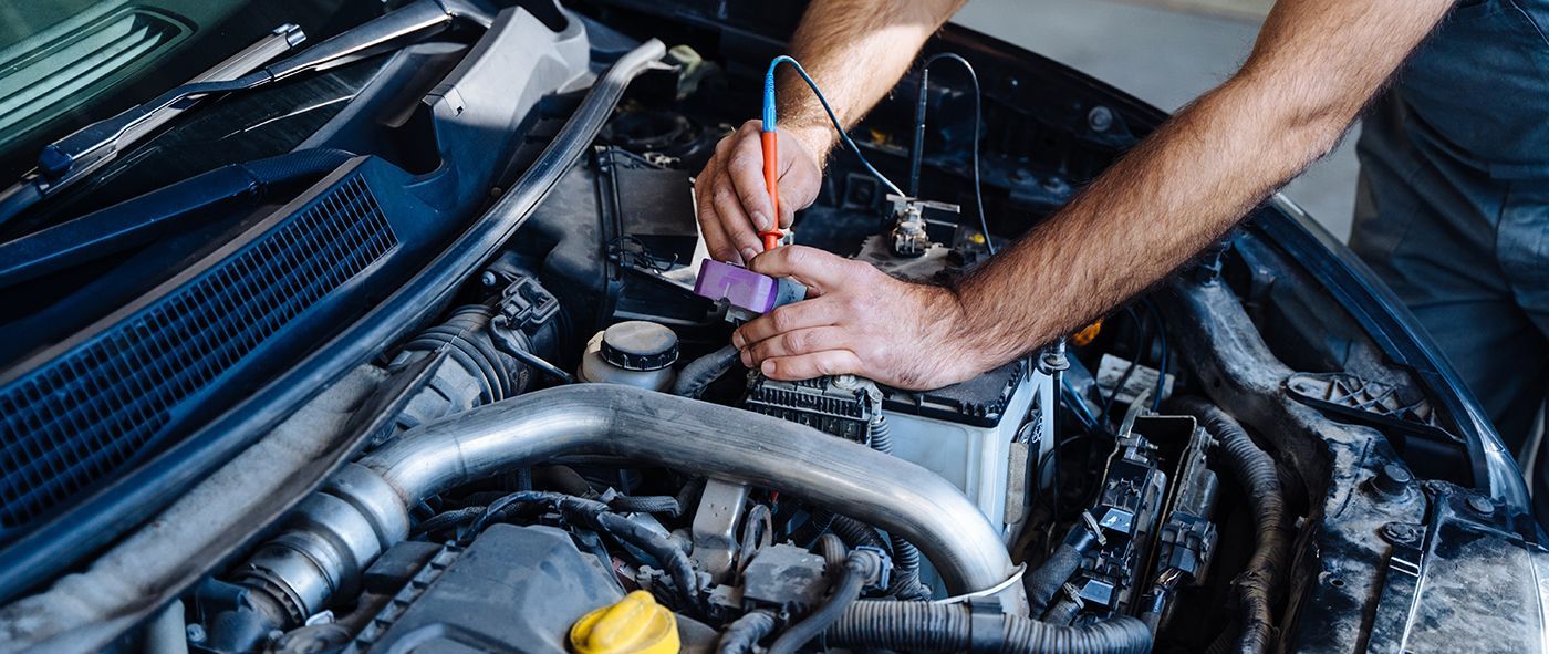 A mechanic tests the battery of a car with a multimeter under the hood of a vehicle.