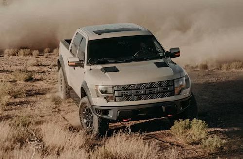 White pickup truck driving off-road through dry grass, kicking up dust.