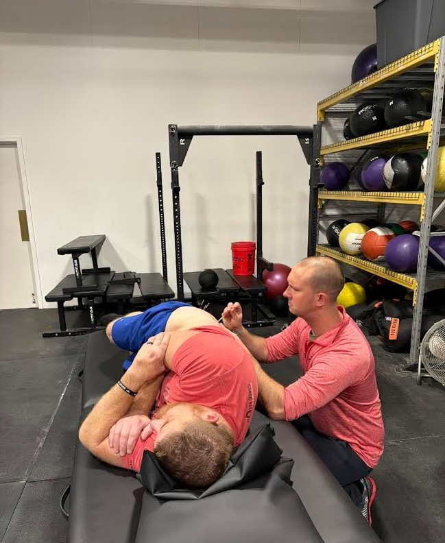 A person receiving shoulder therapy lying on a table while another person assists. A gym setting.