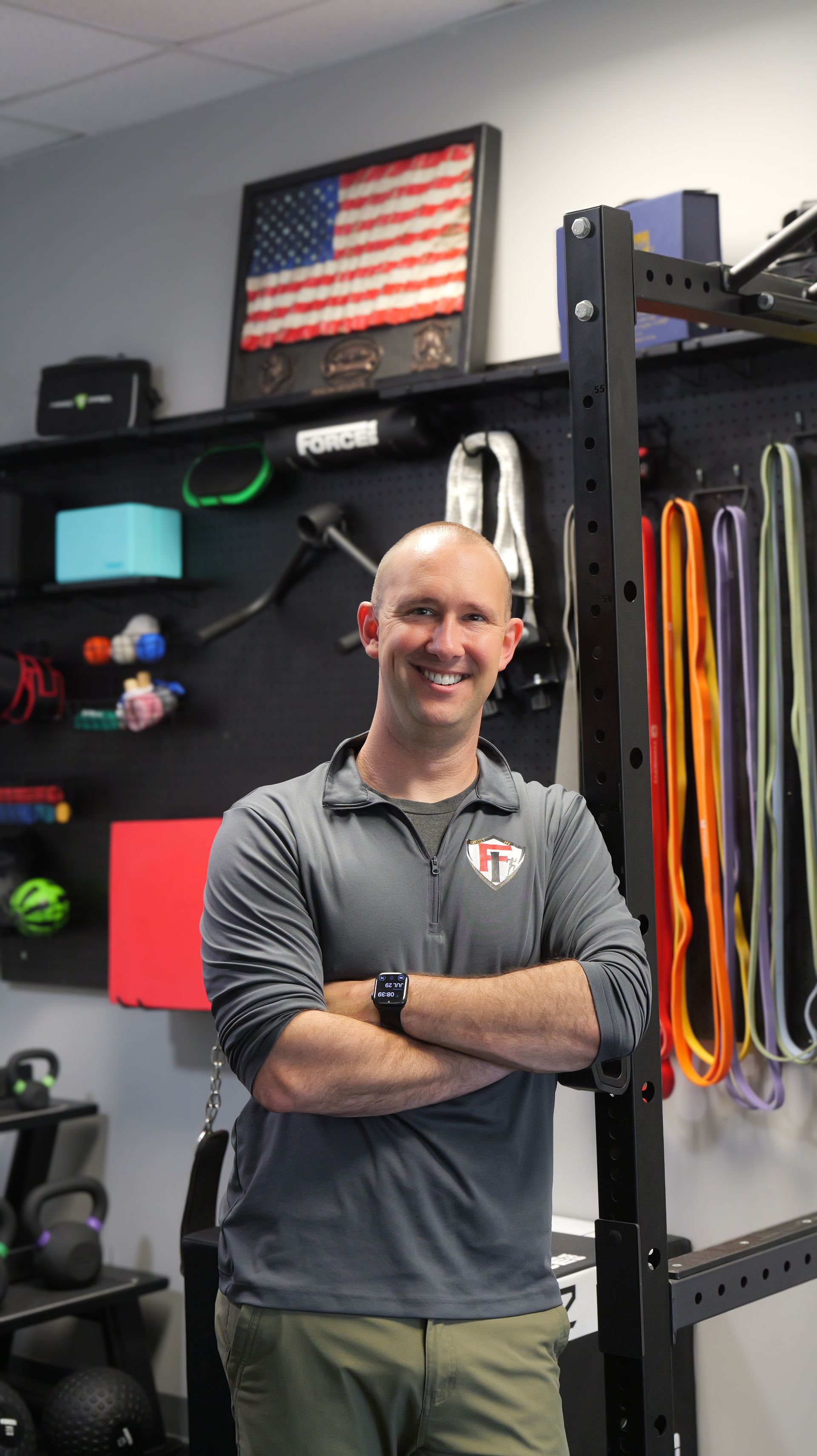 Man in gray shirt with arms crossed, smiling in a gym. Shelves with exercise equipment and American flag in background.