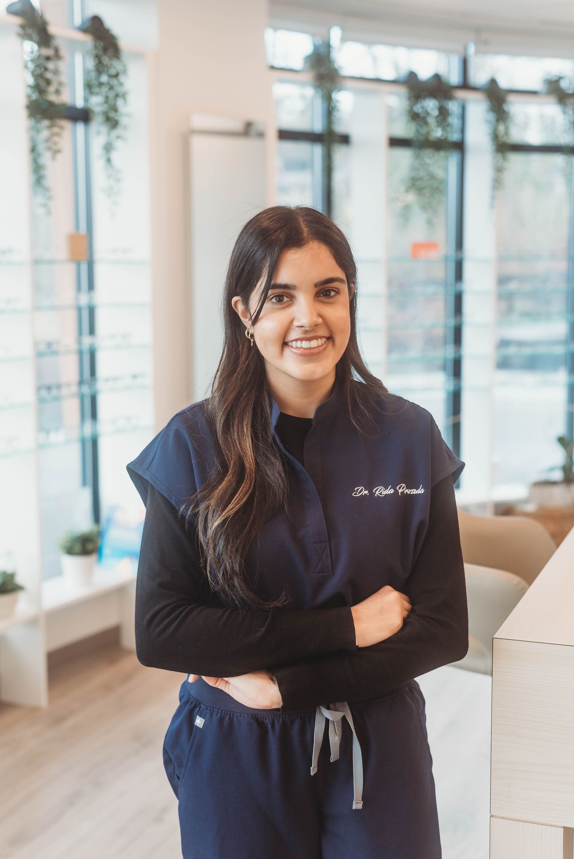 Woman in blue scrubs smiling, arms crossed. Standing in well-lit office, with a window and plants.