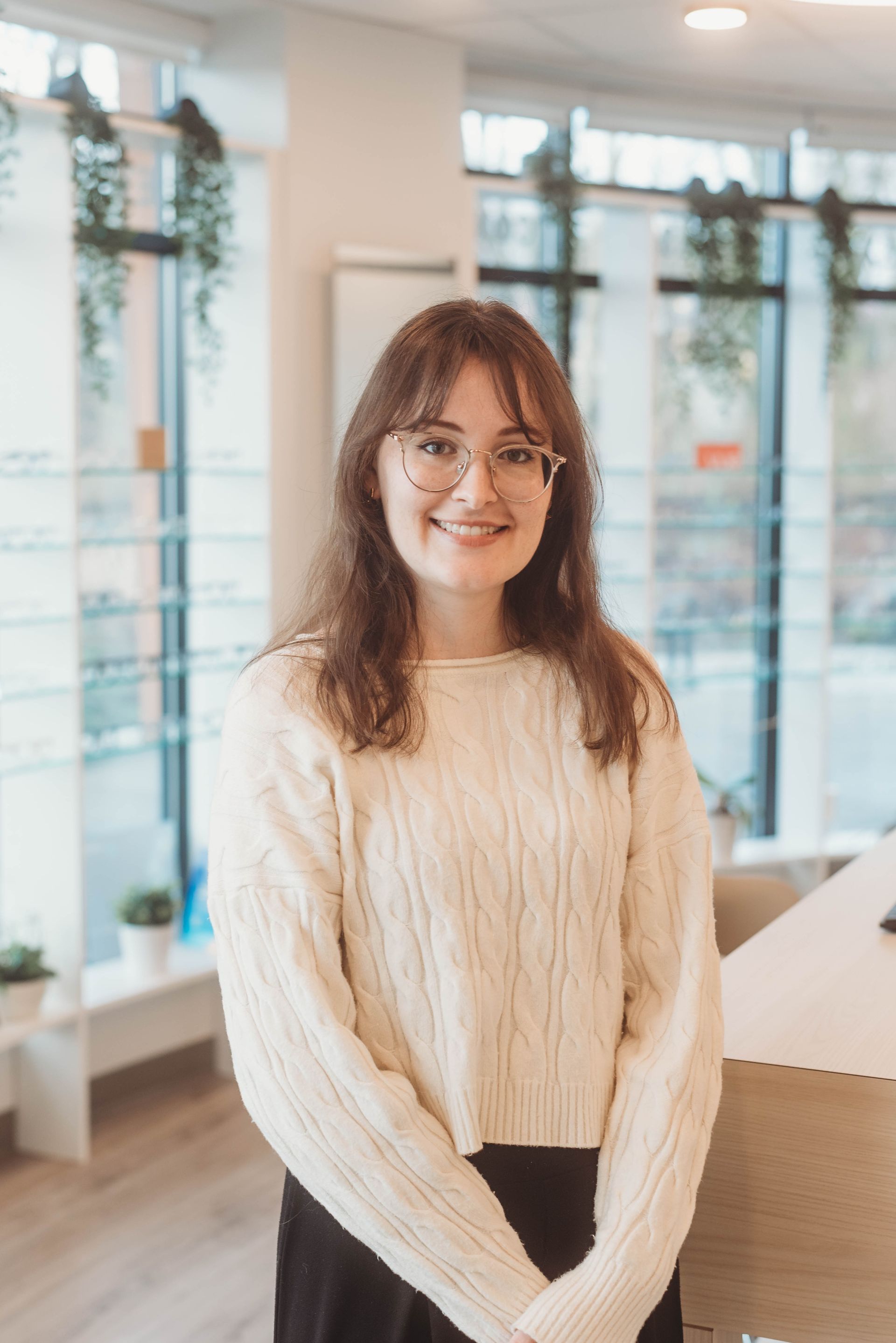 Woman in a cream sweater and glasses smiles at the camera, inside a modern office with greenery.