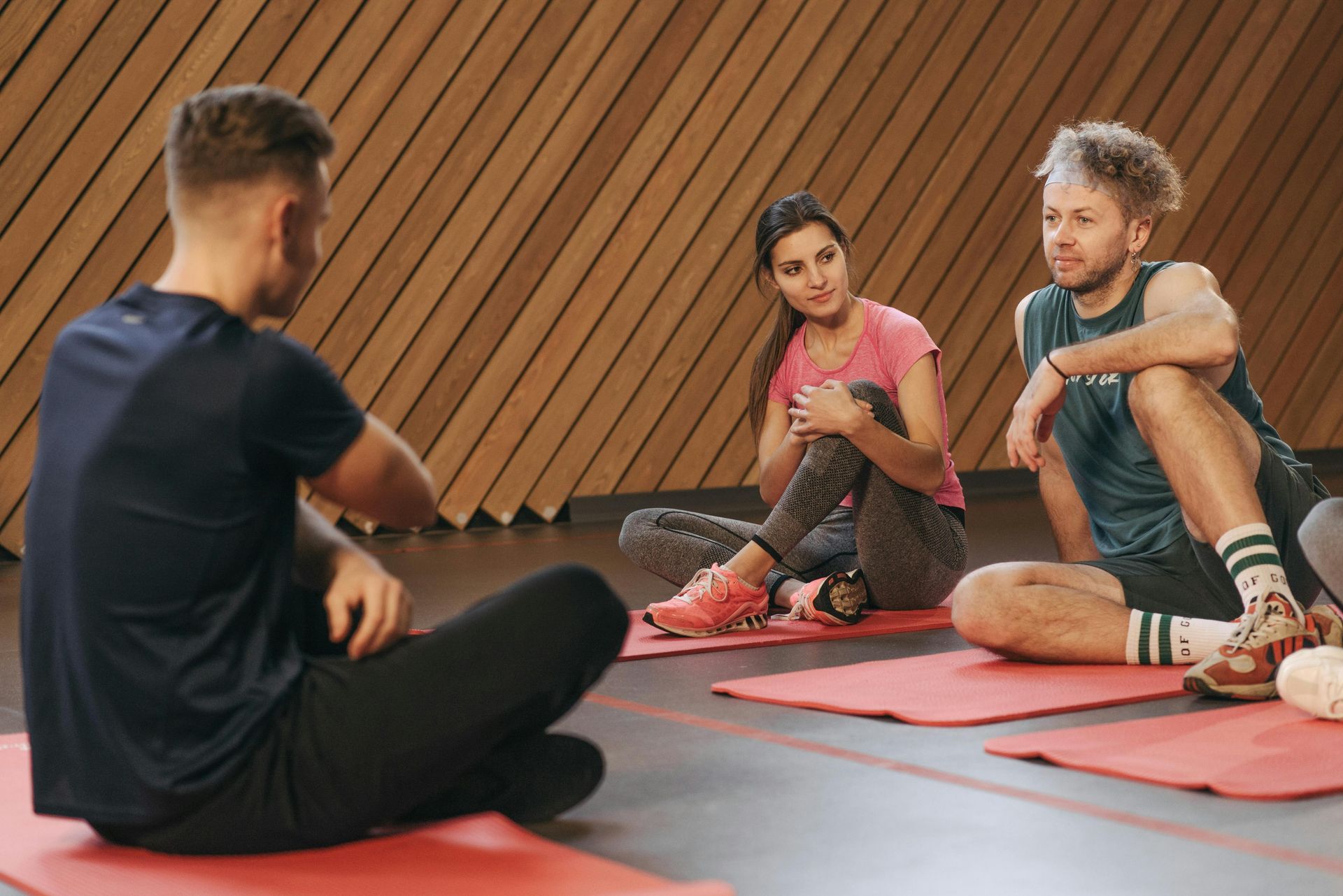 Three people seated on mats, talking. A wooden wall in the background.