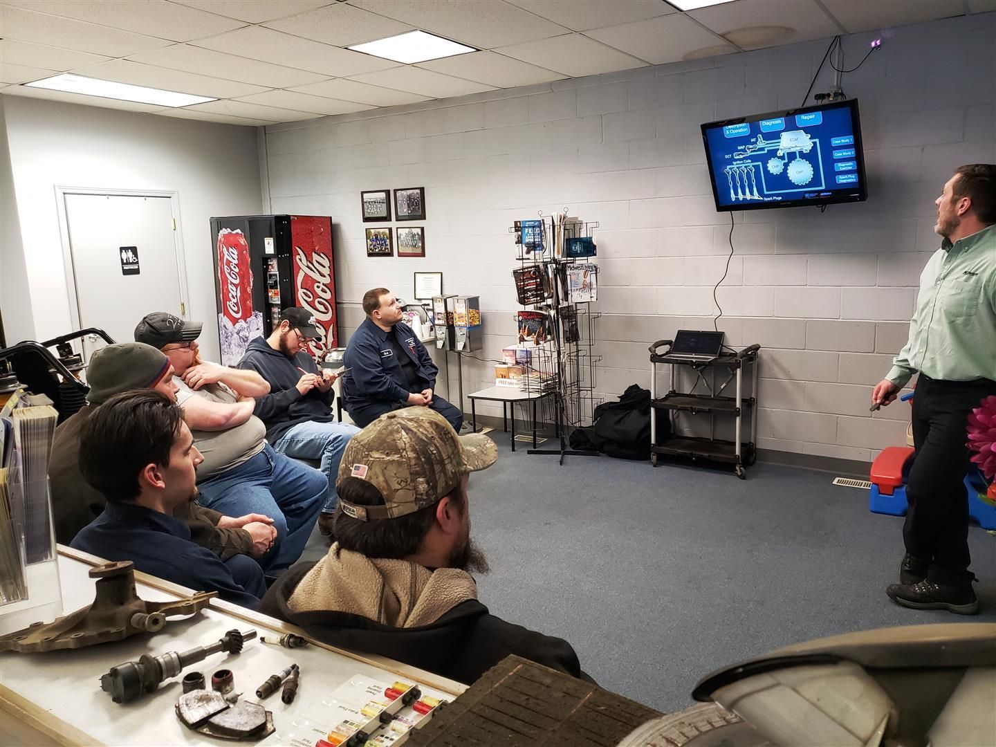 A man in green shirt presents to a group. Attendees seated in a workshop, looking at a screen displaying a diagram. | Quality Car & Truck Repair