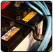 Car battery with attached cables. The battery is black with yellow labels, and blue cables are nearby. | Quality Car & Truck Repair
