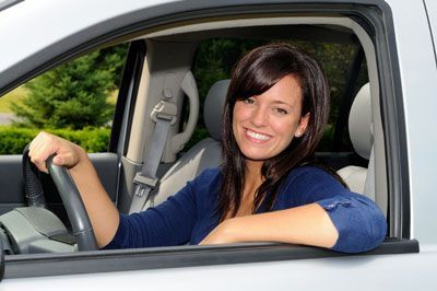 Woman smiling and holding the steering wheel in a silver car, with arm resting on the open window. | Quality Car & Truck Repair Inc