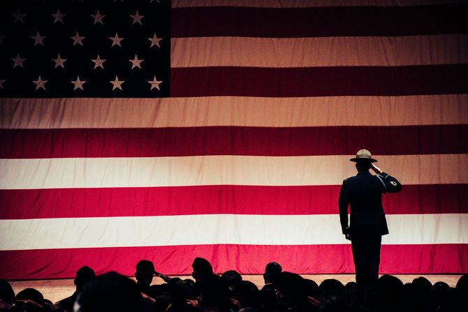 Soldier salutes large American flag; other figures salute in the foreground, indoors.