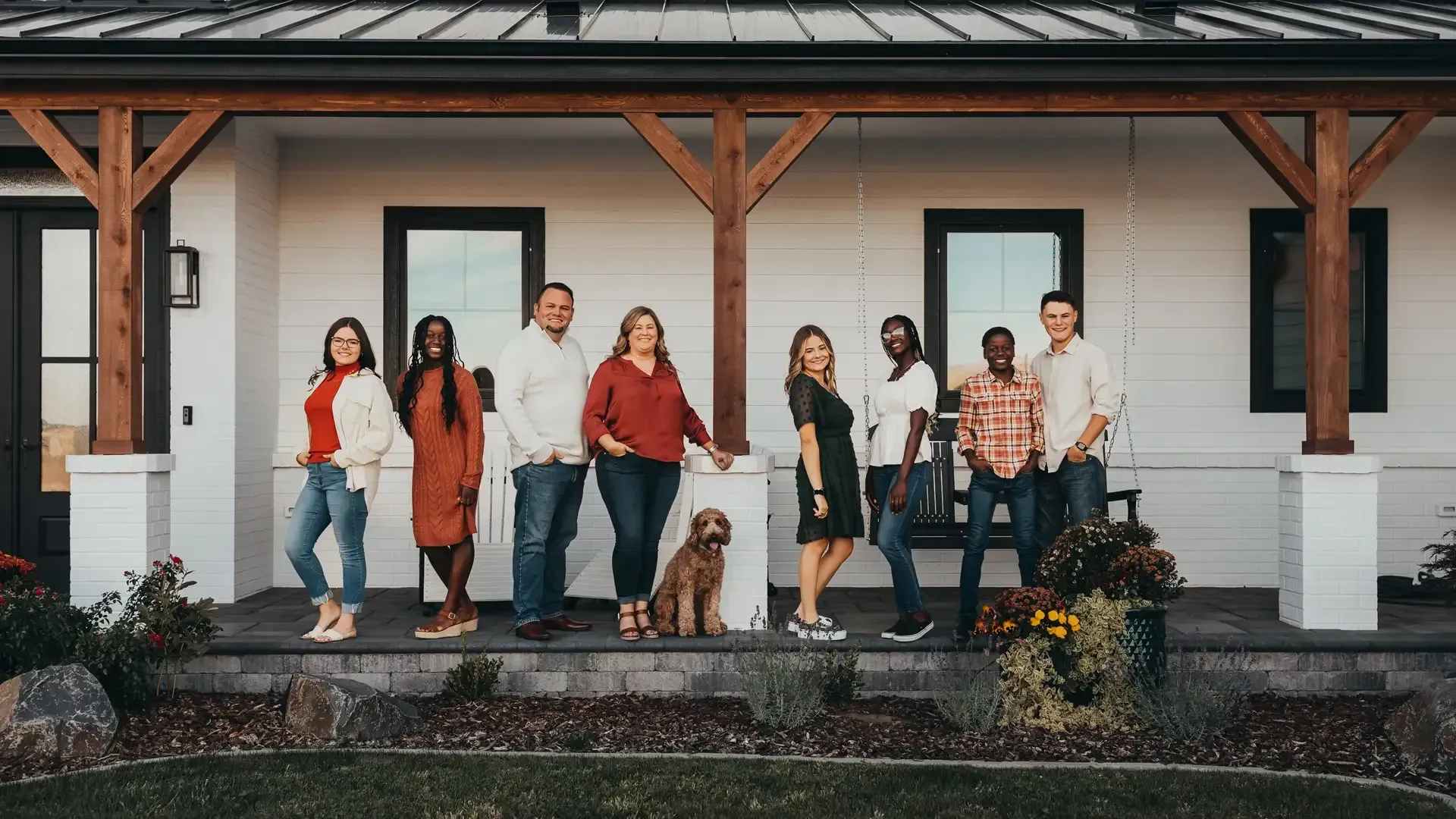 Group of people standing on a porch in front of a white house; a dog sits in front.