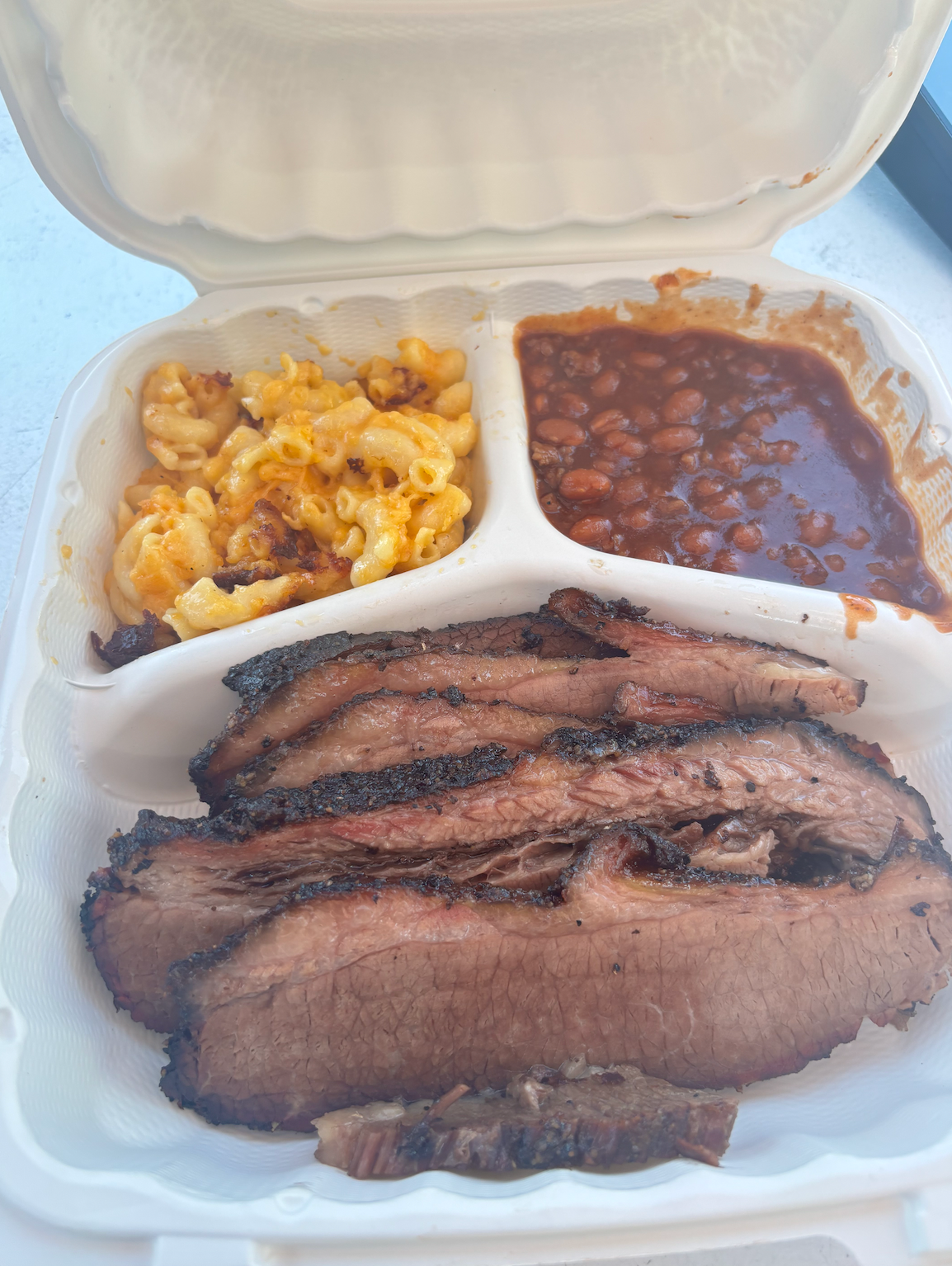A styrofoam container filled with meat , macaroni and cheese , and beans.
