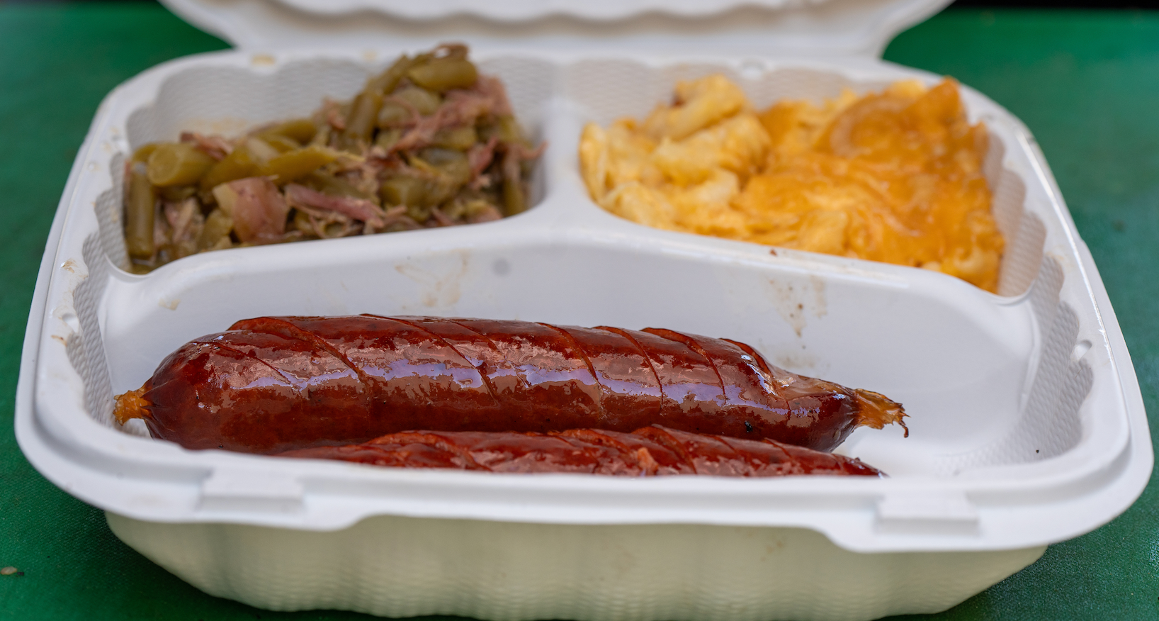 A styrofoam container filled with a sausage , macaroni and cheese , and green beans.