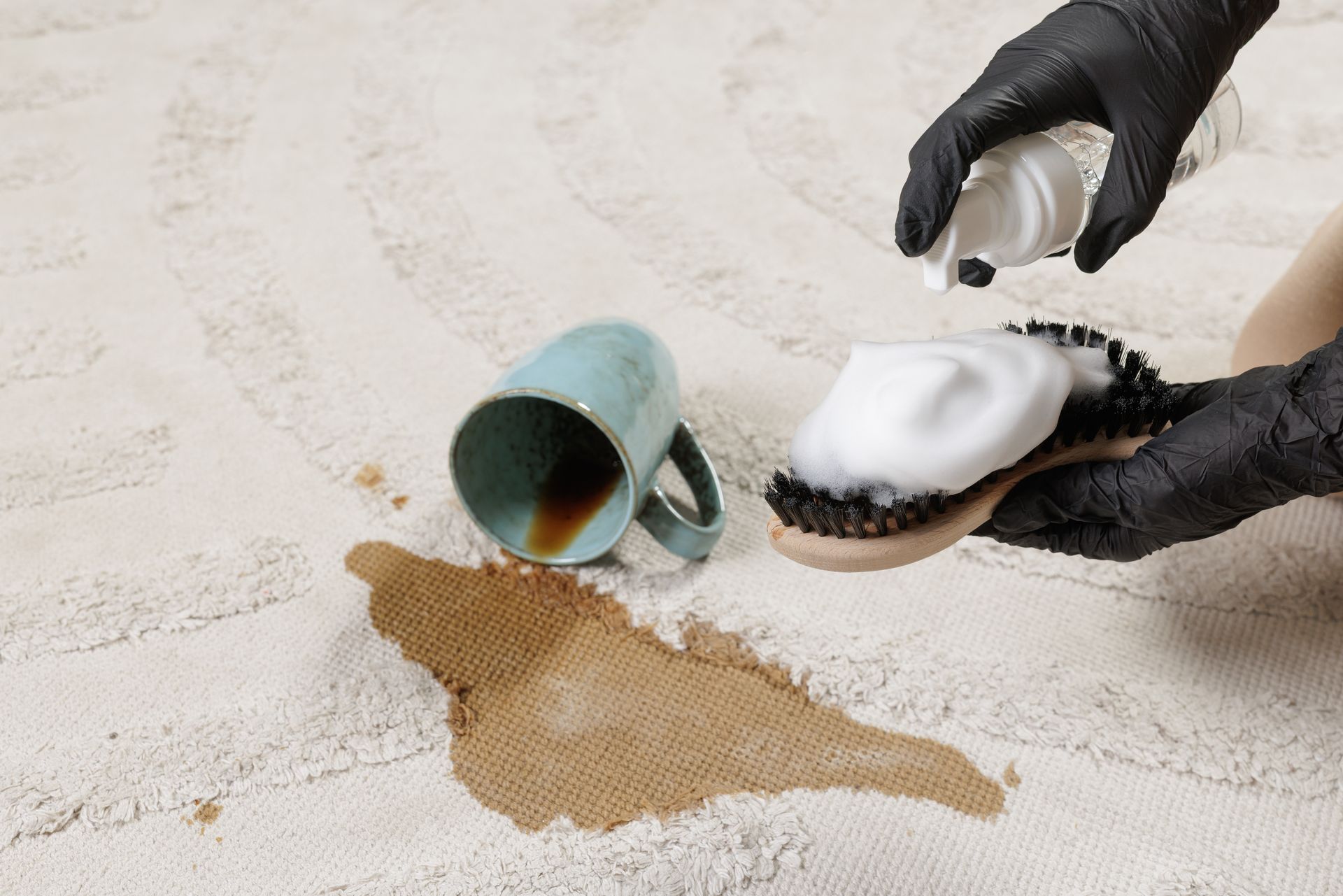 Gloved hands cleaning a coffee spill from a white carpet beside an overturned mug