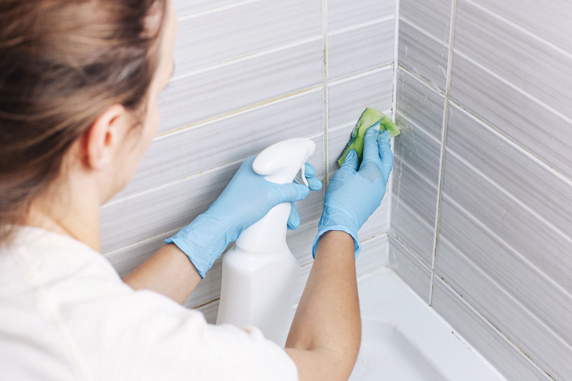 Person in blue gloves cleaning bathroom tile grout with a spray bottle and cloth