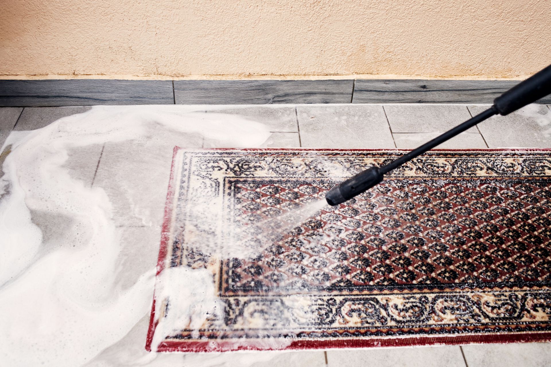 Pressure washing a patterned rug on a tiled patio with white suds and spray