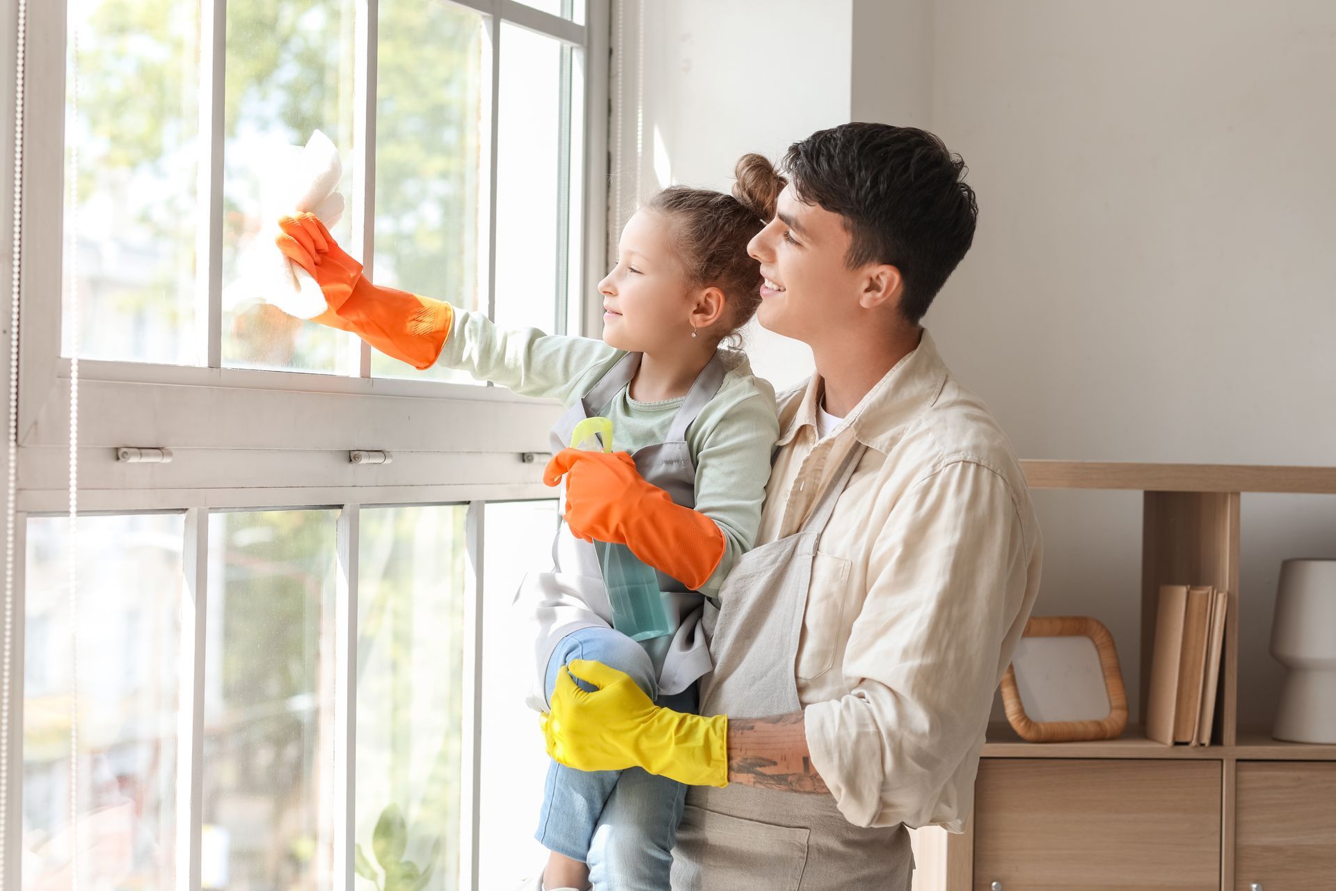 Adult and child cleaning a window indoors, both wearing bright rubber gloves.