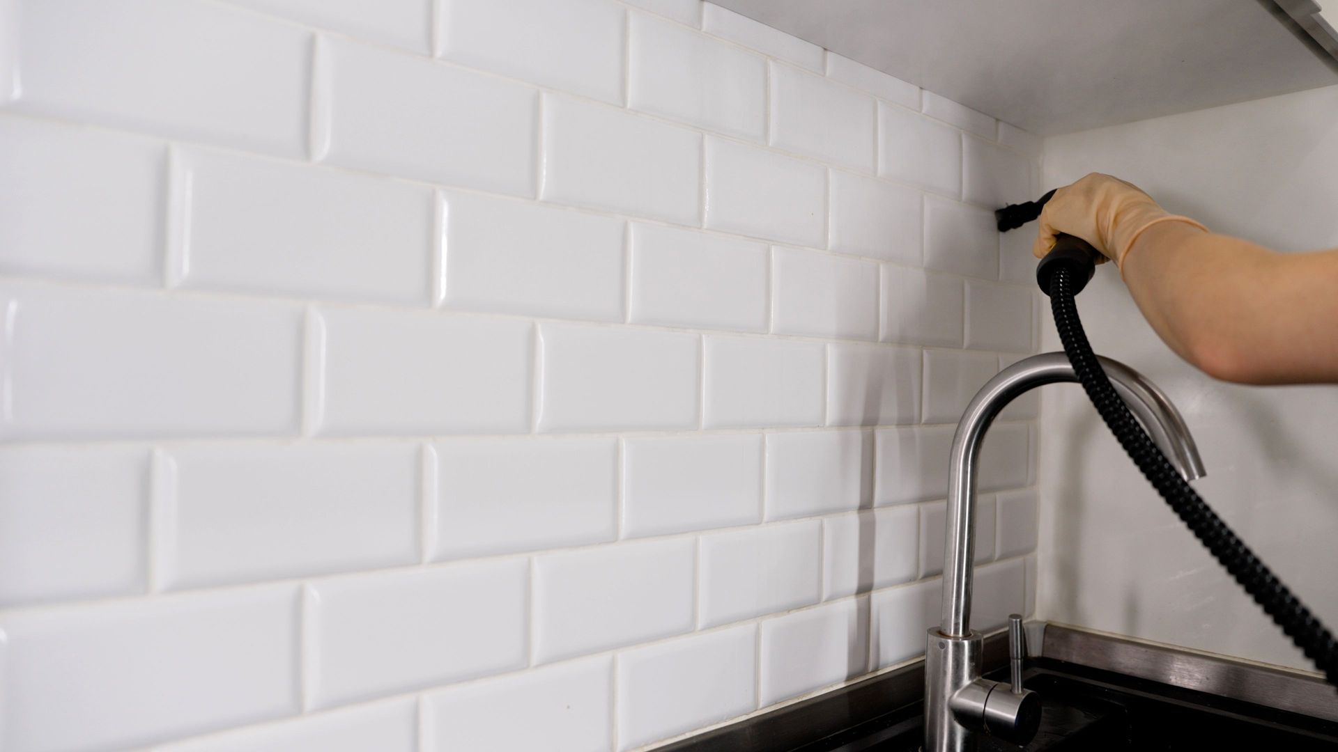 Hand cleaning white subway tile backsplash with a black spray nozzle over a kitchen sink