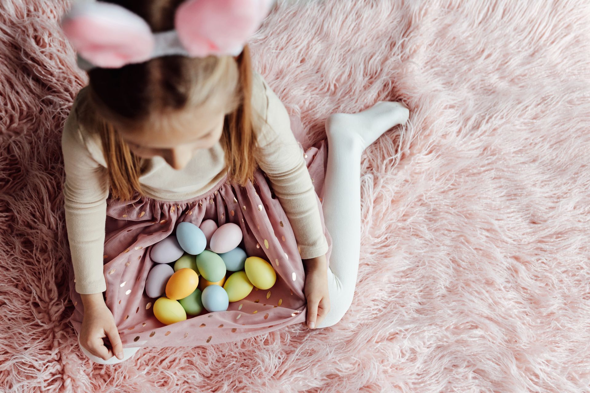 Girl wearing bunny ears, surrounded by pink fur and pastel Easter eggs in her skirt.