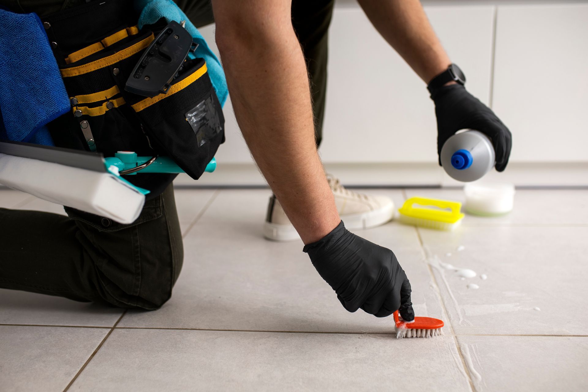 Person wearing gloves scrubbing tile grout with a brush, cleaning supplies nearby.