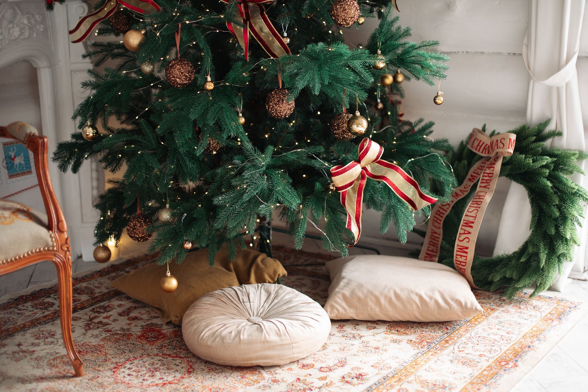 Christmas tree with ornaments and bows, surrounded by pillows on a rug near a window.