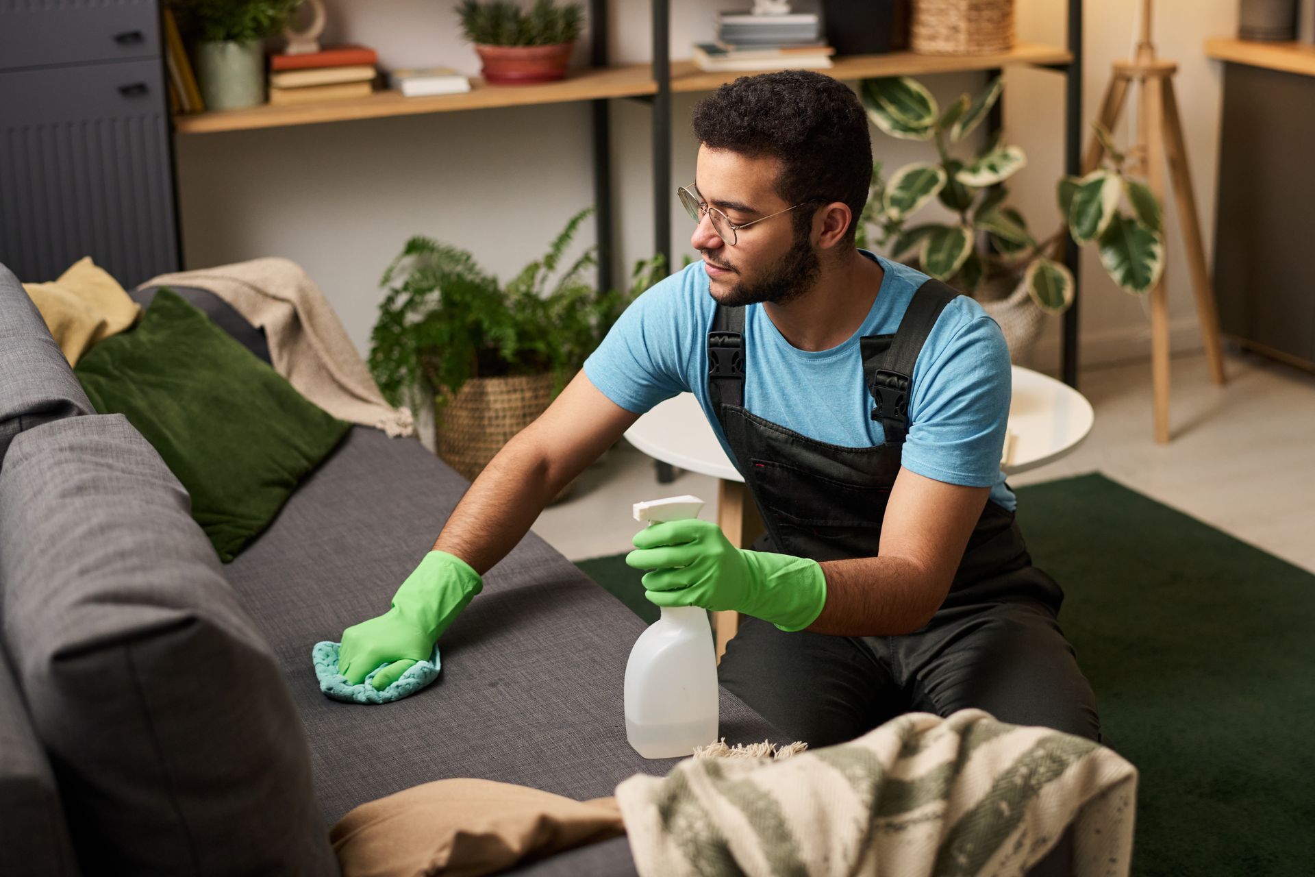Person wearing gloves cleans gray couch with spray bottle in a living room.