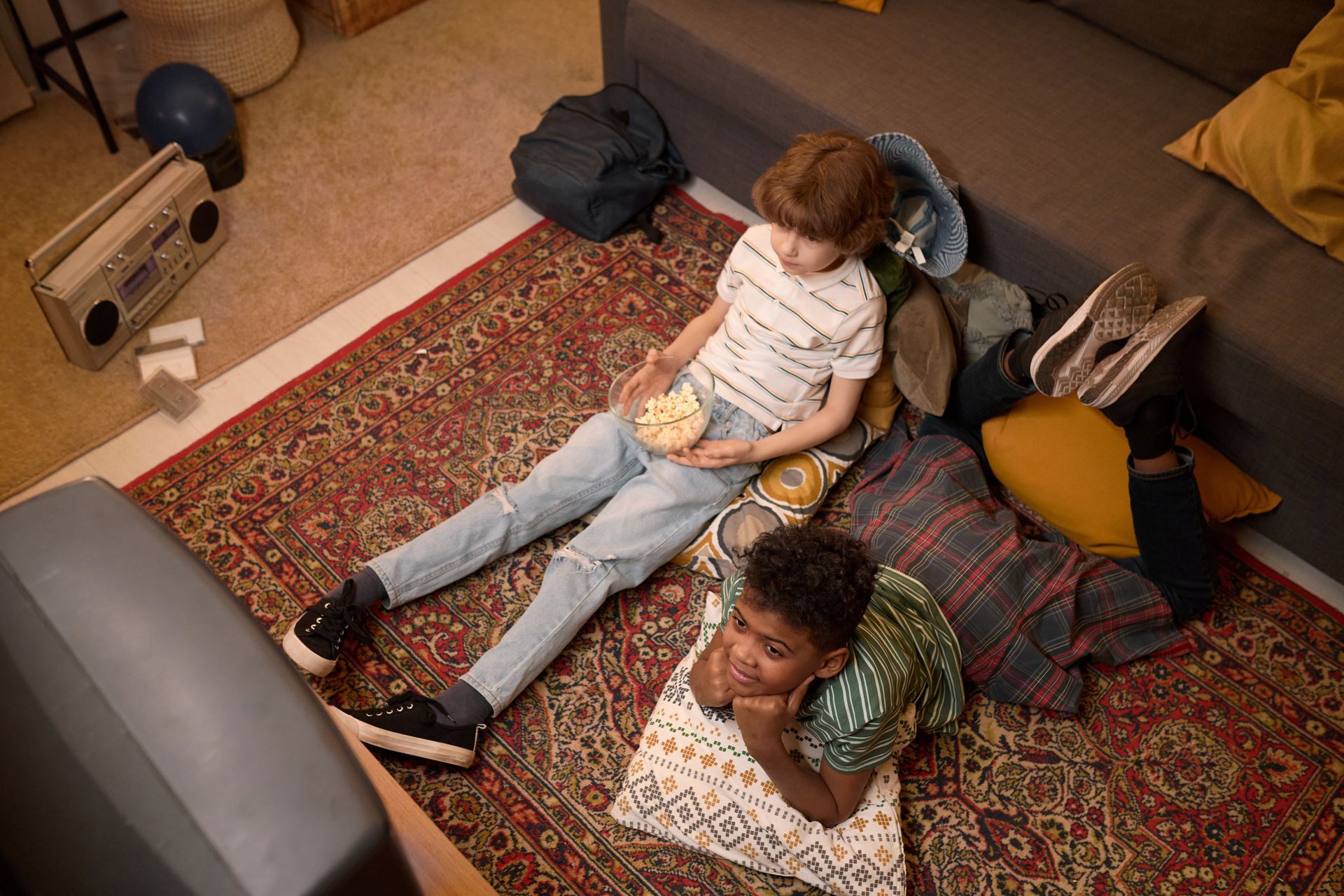 Two children watching TV, one holding popcorn, lounging on a patterned rug near a couch.