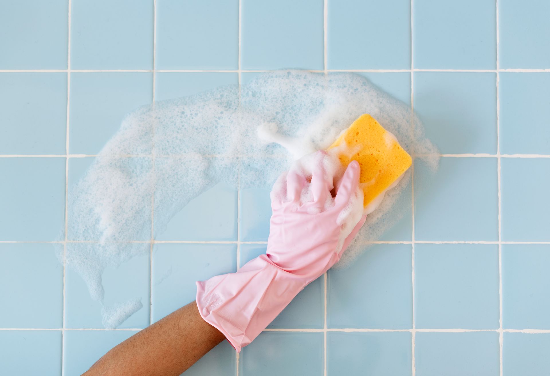 Hand in pink glove scrubbing light blue tiled wall with yellow sponge and soap.