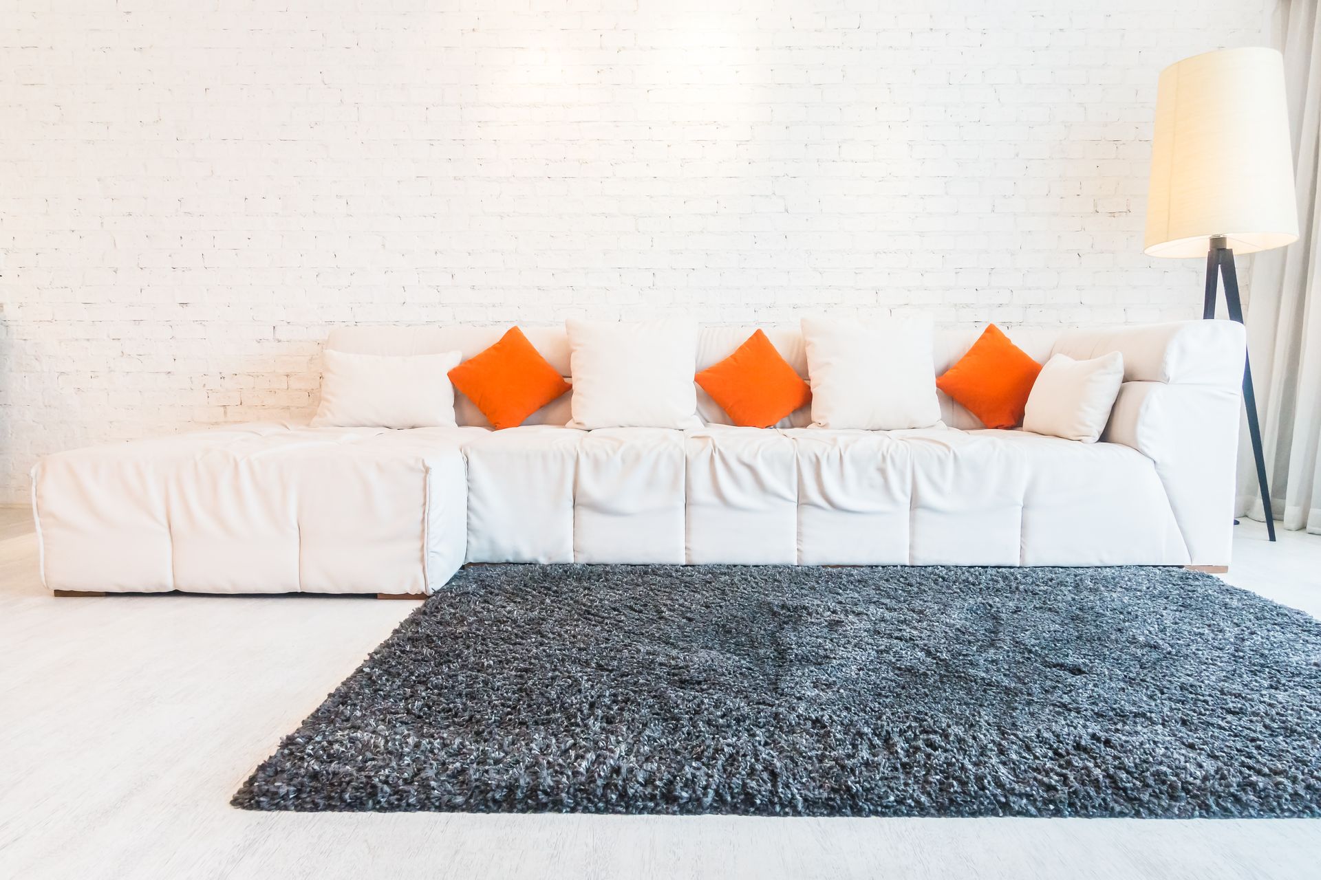 White sofa with orange pillows, gray rug, and lamp against a white brick wall.