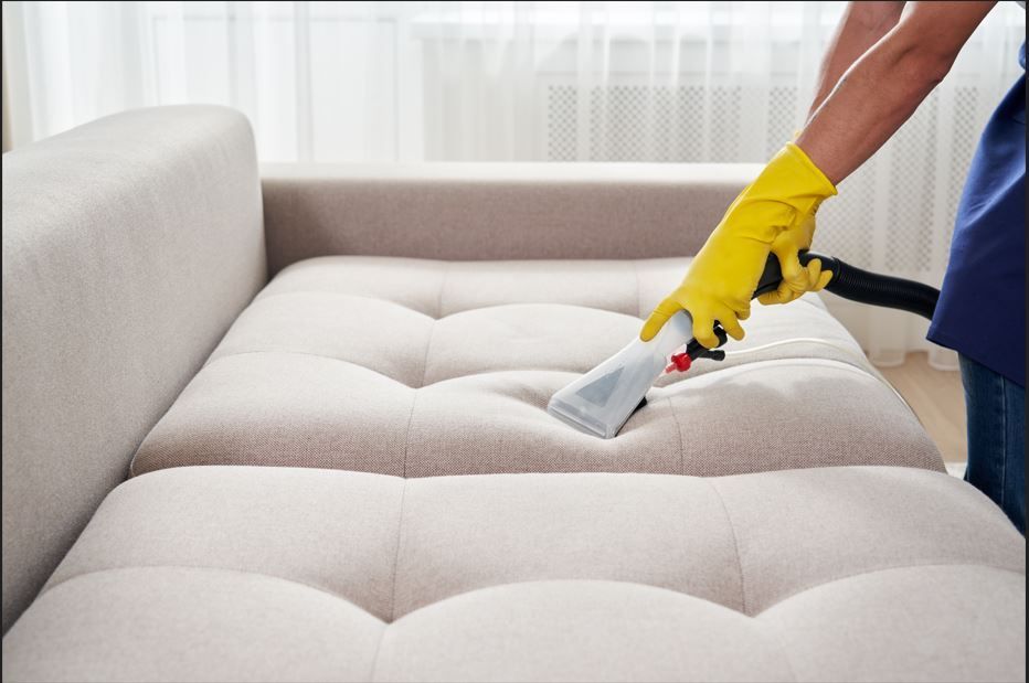 Person in yellow gloves cleaning a beige tufted couch with a handheld upholstery cleaner.