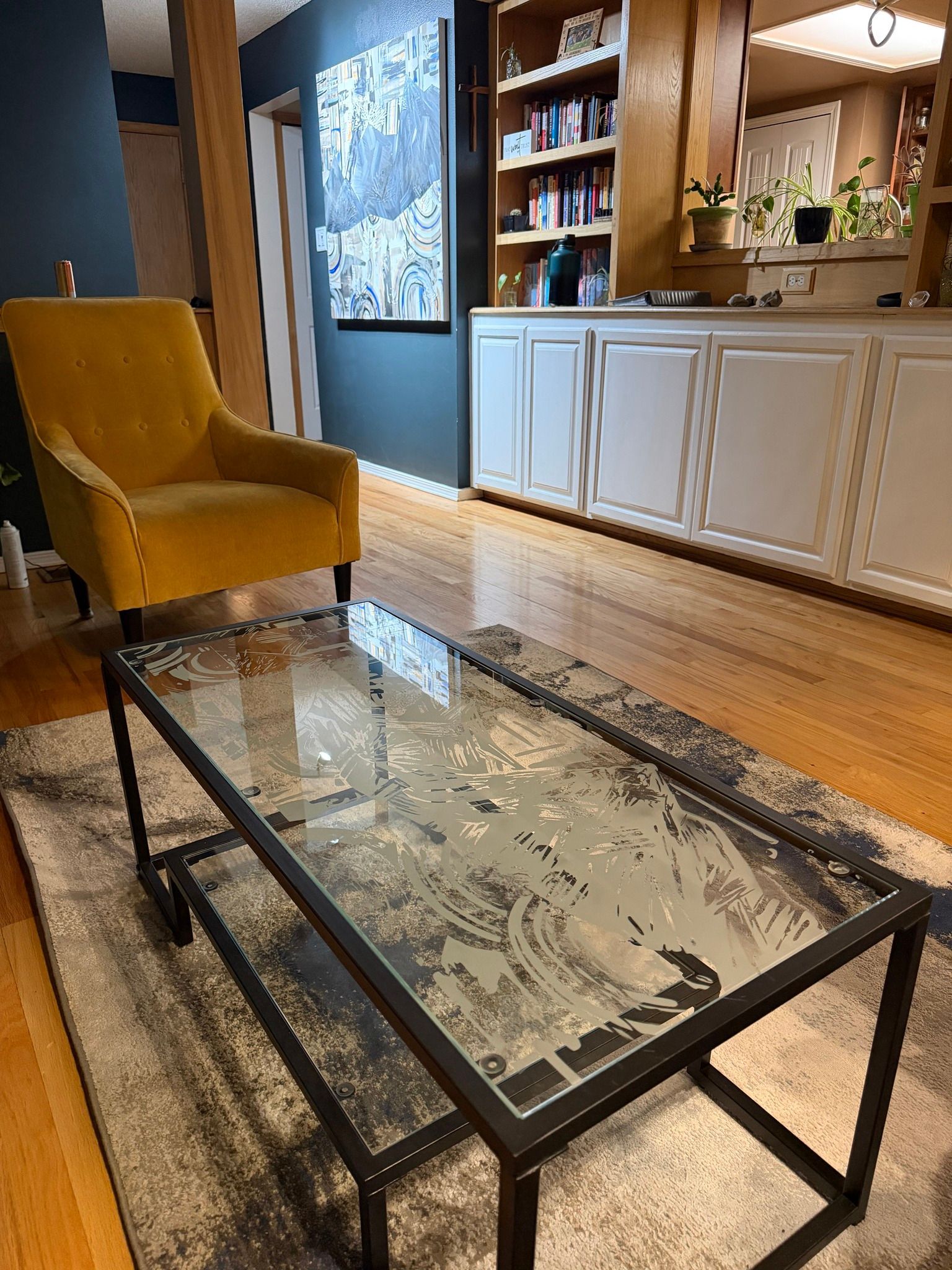 Living room with mustard armchair, glass coffee table, wood floor, and built-in white cabinets and shelves.