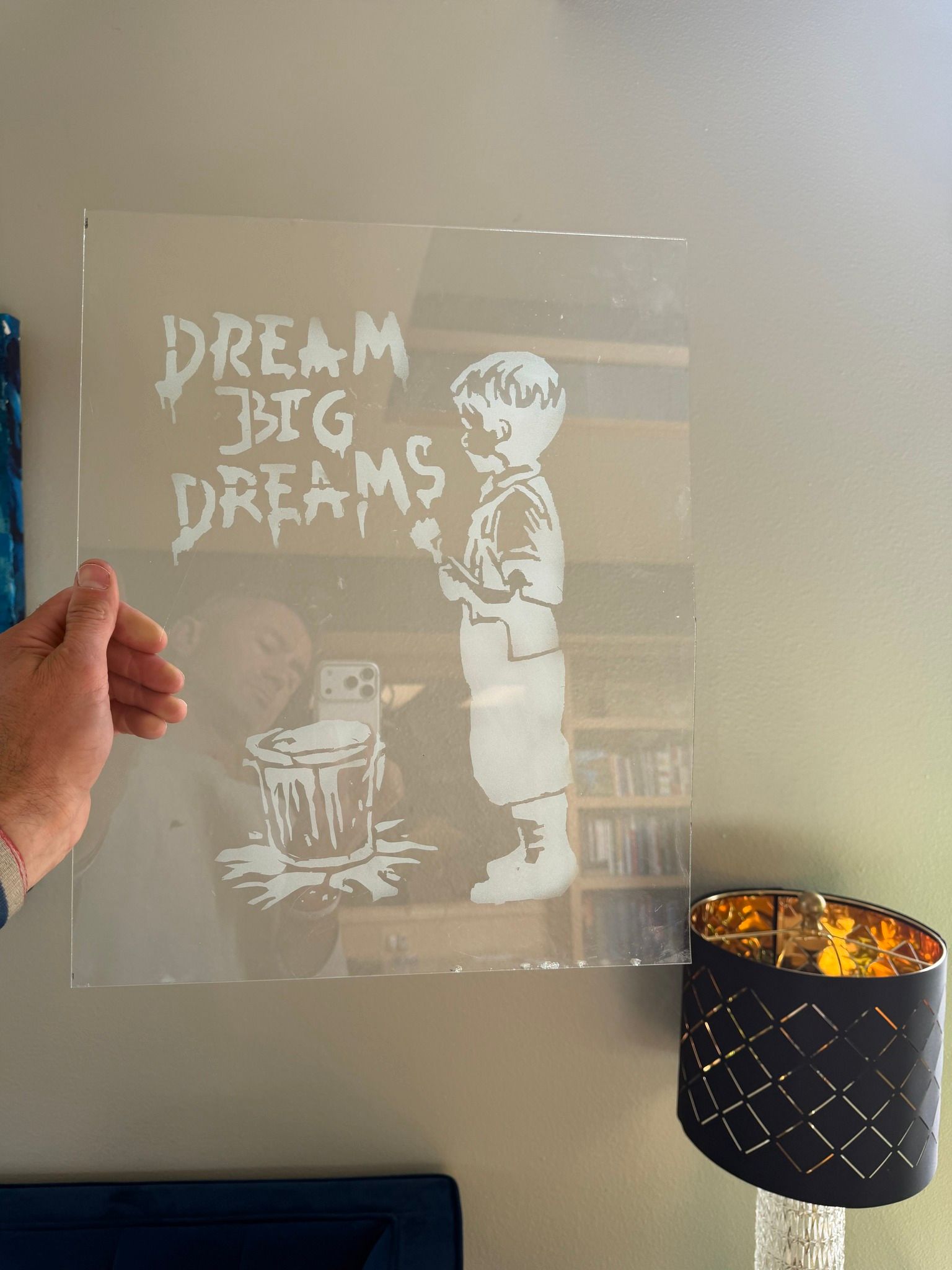 Hand holding a translucent poster reading “DREAM BIG DREAMS” beside a lit table lamp on a wall.