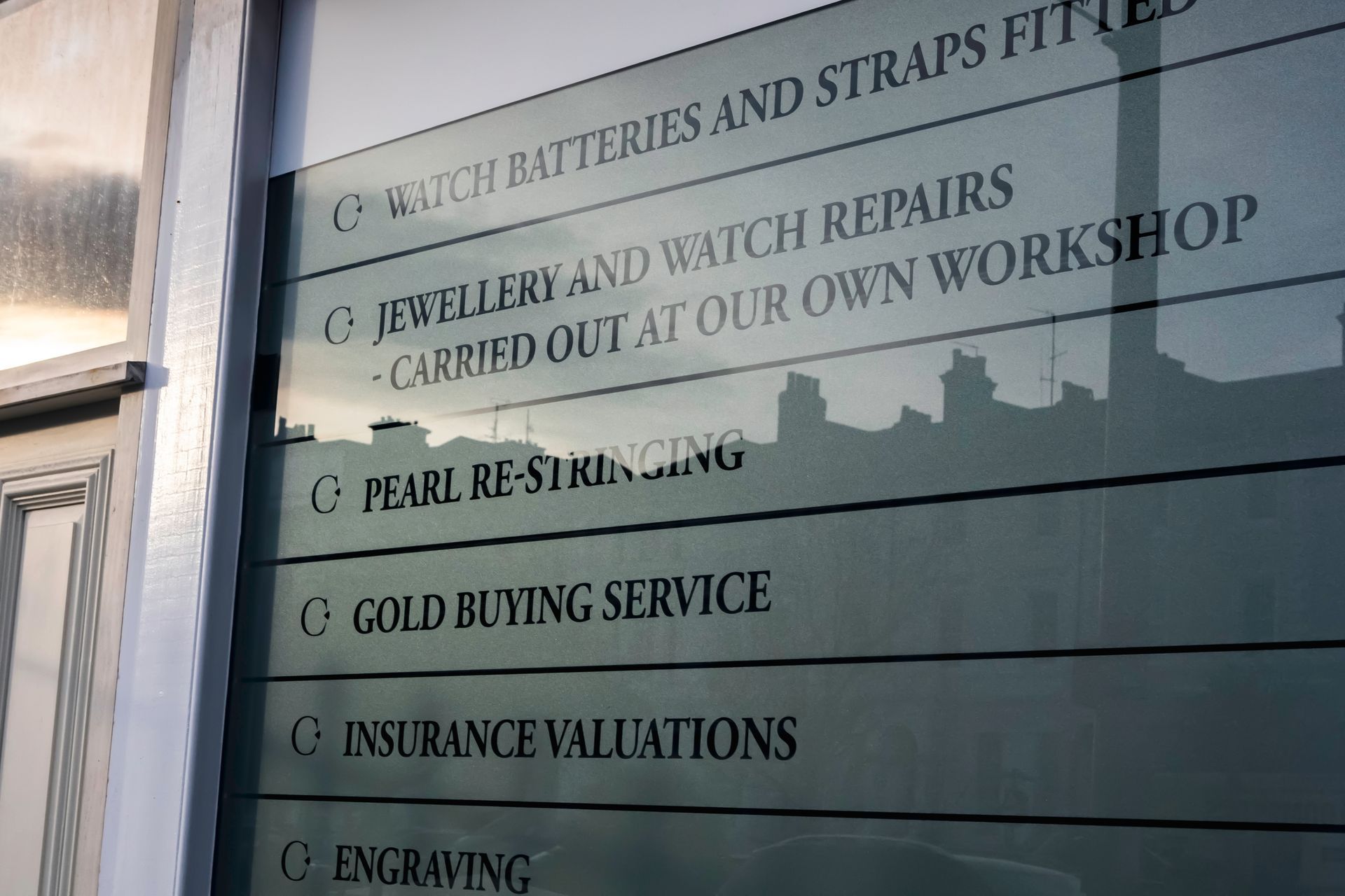 Storefront window with a gold-lettered list of jewelry and watch repair services, including engraving and valuations