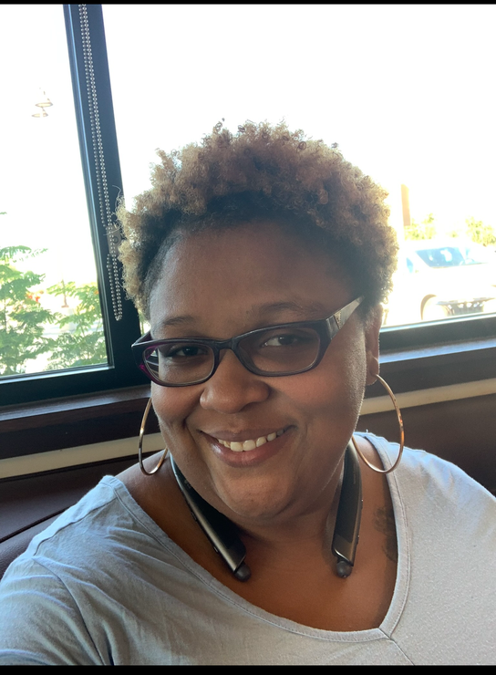 Person smiling, wearing glasses, large hoop earrings, and a light gray shirt, seated near a window.