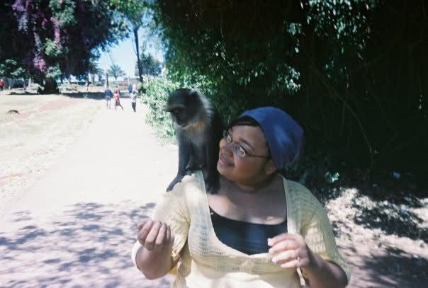 Woman with a monkey on her shoulder smiles, outdoors on a sunny path.