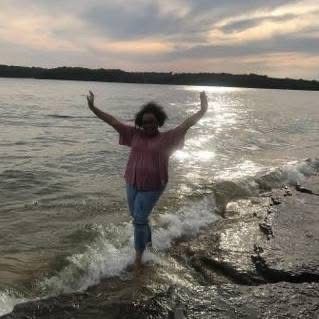 Person with arms raised standing in water by a shoreline, with sunlit water and sunset sky.