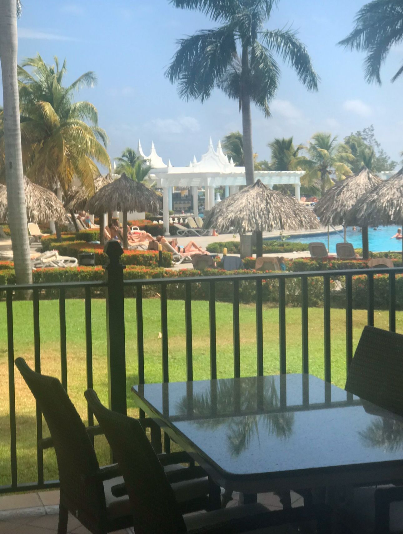 Patio view of a resort pool area with palm trees, thatched umbrellas, and white gazebo.