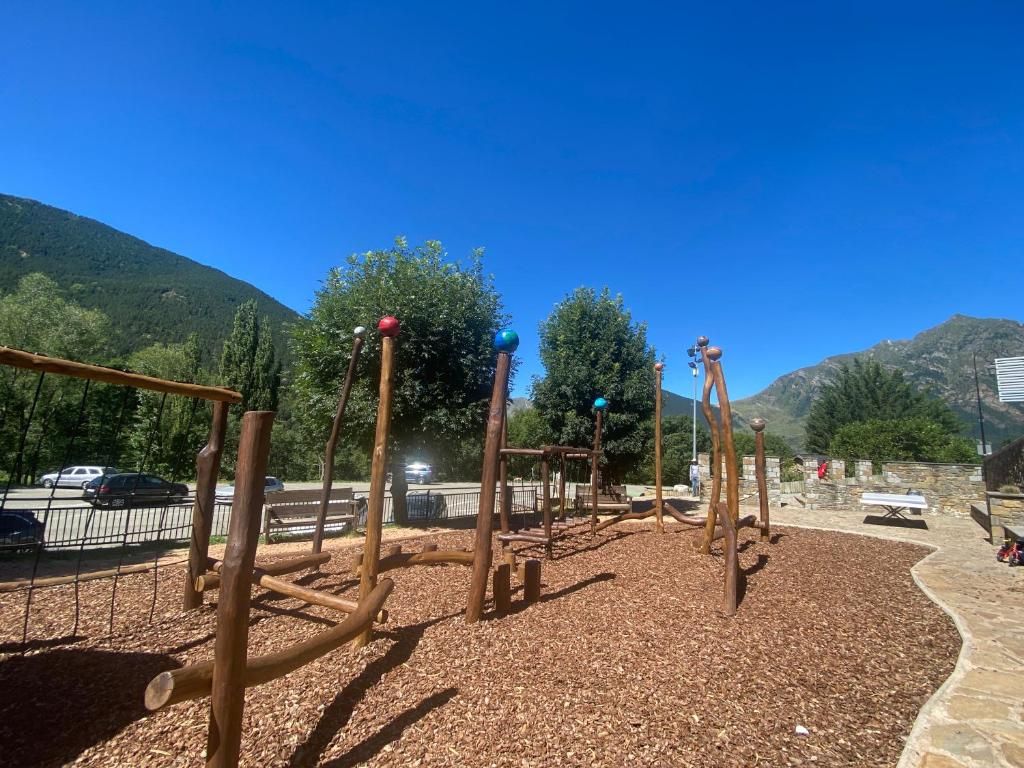 Parc infantil amb estructures de fusta sobre terra d'estelles de fusta, arbres i muntanyes sota un cel blau.